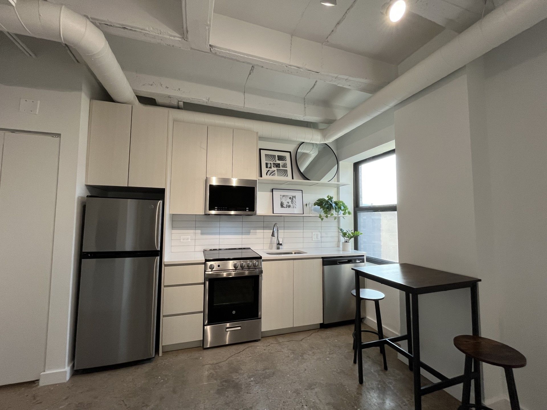 Small kitchen with stainless steel appliances and light cabinets. A small black table with two stools sits by a window.