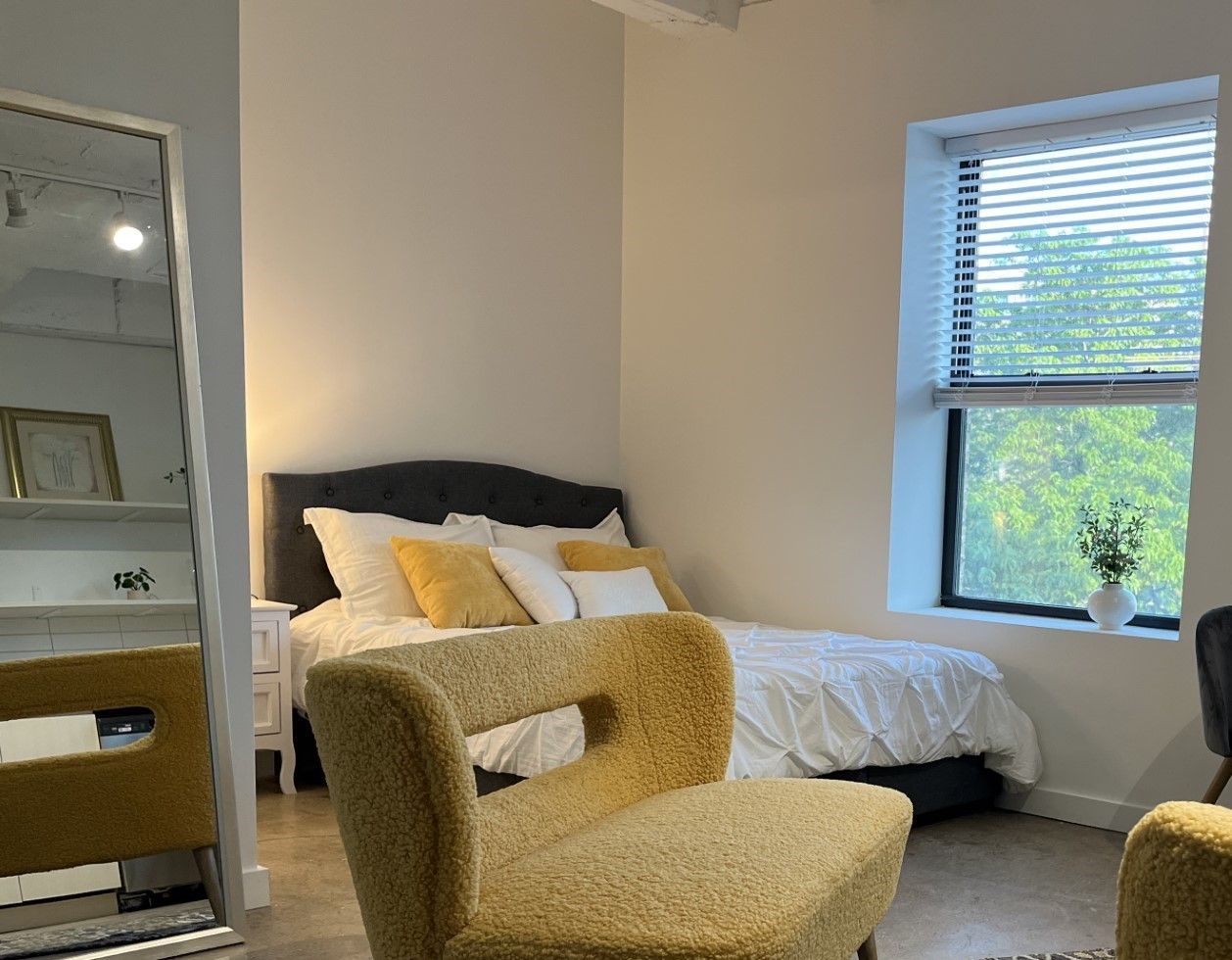 Bedroom with a yellow chair in the foreground, bed with yellow pillows, window with blinds, and a full-length mirror.