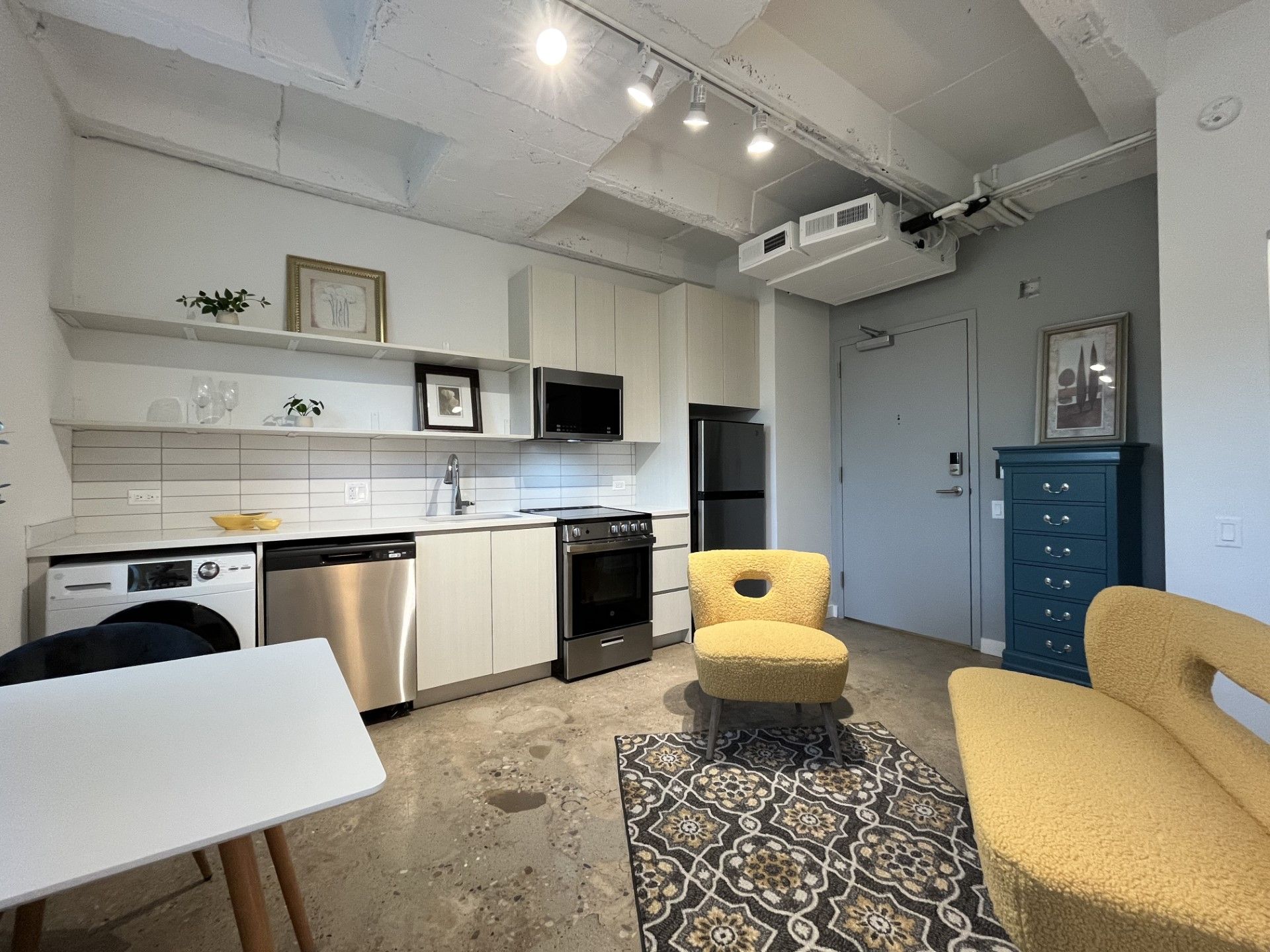 A small, modern apartment kitchen and living space. Light-colored cabinetry and walls, stainless steel appliances, and a yellow armchair.