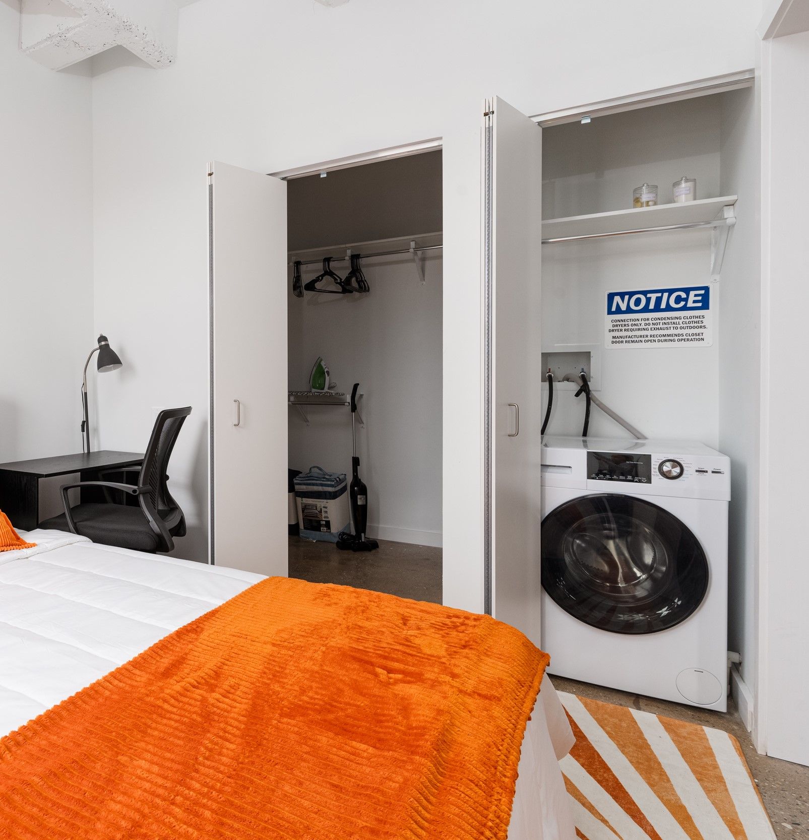 A bedroom with a washing machine and closet. White walls and closet doors frame the washer. An orange blanket adorns the bed.