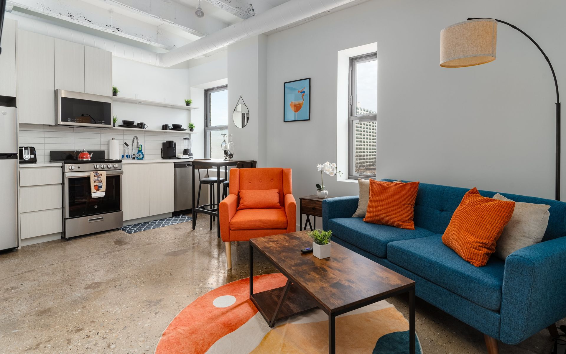 Modern apartment interior with a blue sofa, orange armchair, and small kitchen. Concrete floor, white walls, and large windows.