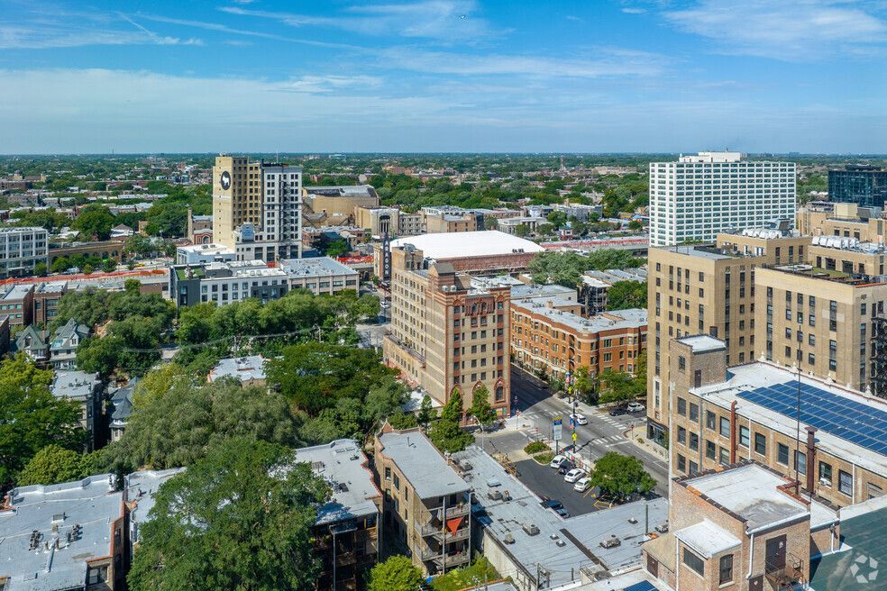 Aerial view of a city with various buildings, including a tall beige tower. Lush green trees and blue sky are also visible.