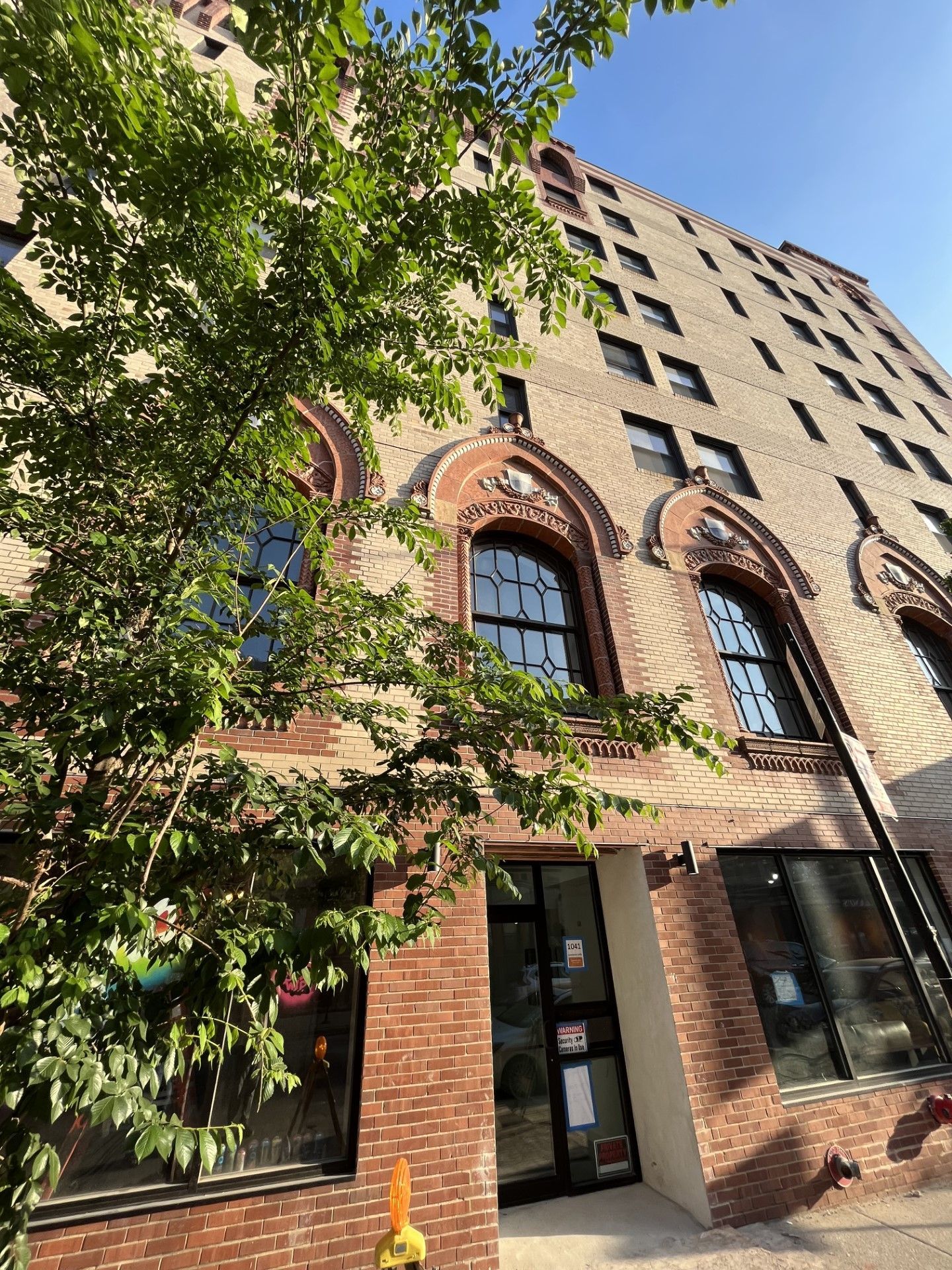 Low-angle shot of a brick building with arched windows and a dark doorway. A tree with green leaves partially obscures the view.