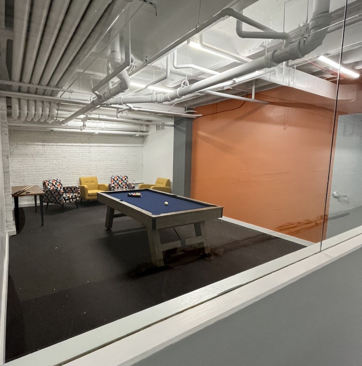 Pool table in a rec room with seating, orange accent wall, and exposed pipes on the ceiling.