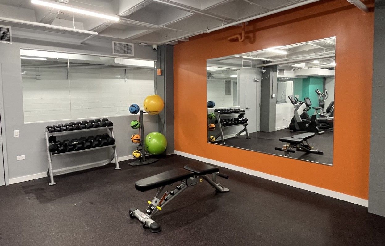 Gym interior with weights, bench, mirrors, and exercise equipment; walls are orange and gray.