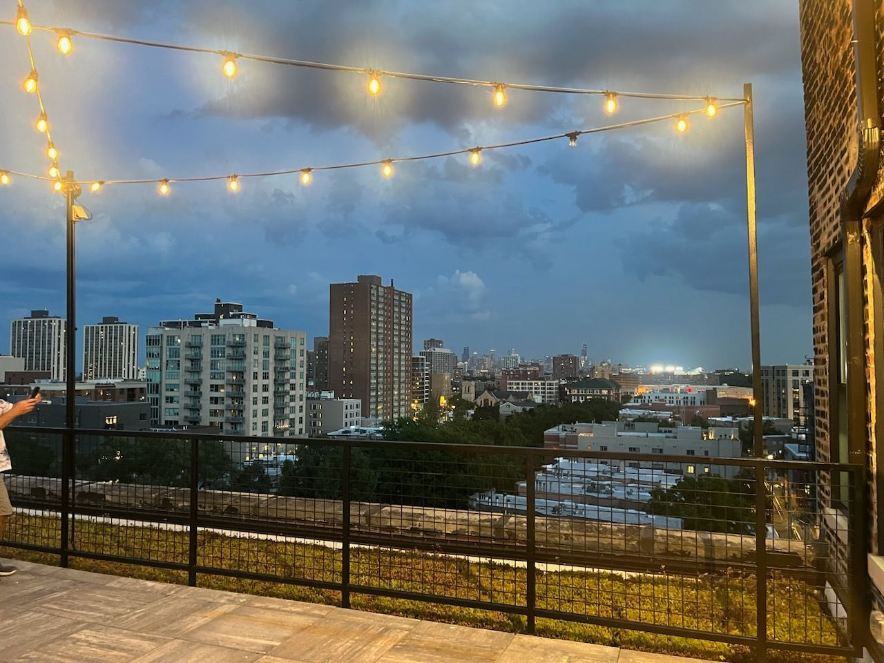 Rooftop patio with string lights, overlooking a city skyline under a cloudy, darkening sky.