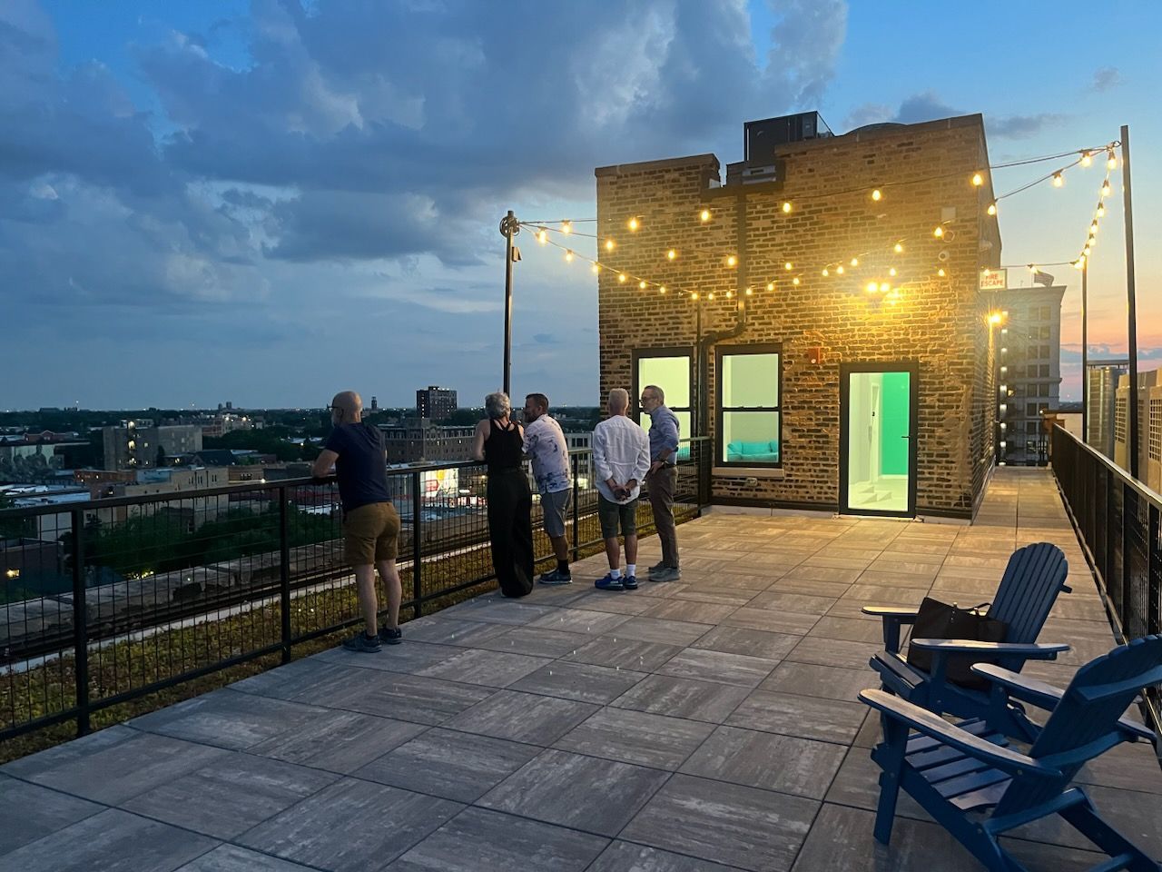 People gather on a rooftop patio at dusk. String lights illuminate a brick building, and cityscape is visible in the distance.