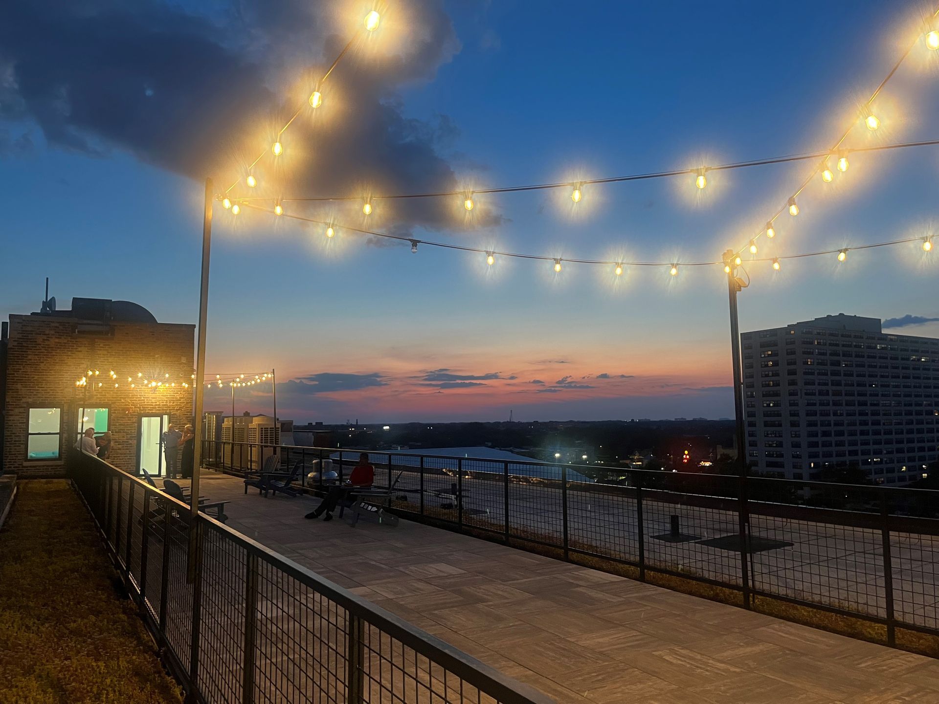 Rooftop patio at dusk with string lights, people seated, and a view of the city skyline against a colorful sky.