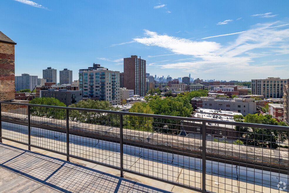View from a rooftop terrace overlooking a cityscape with various buildings under a bright blue sky. A metal railing and the side of a brick building are in the foreground.