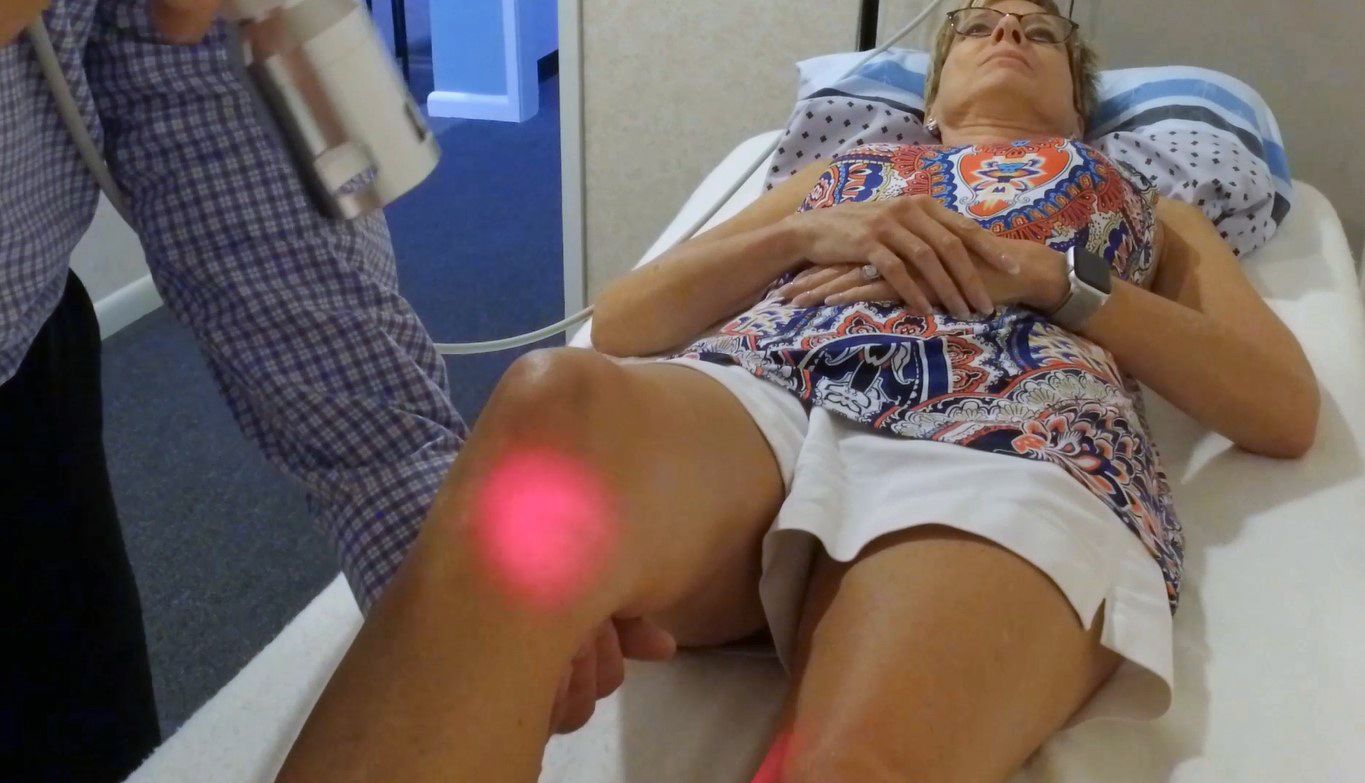 A person receives laser therapy on their knee while lying on an examination table. A red light is visible in Fayetteville, NC.