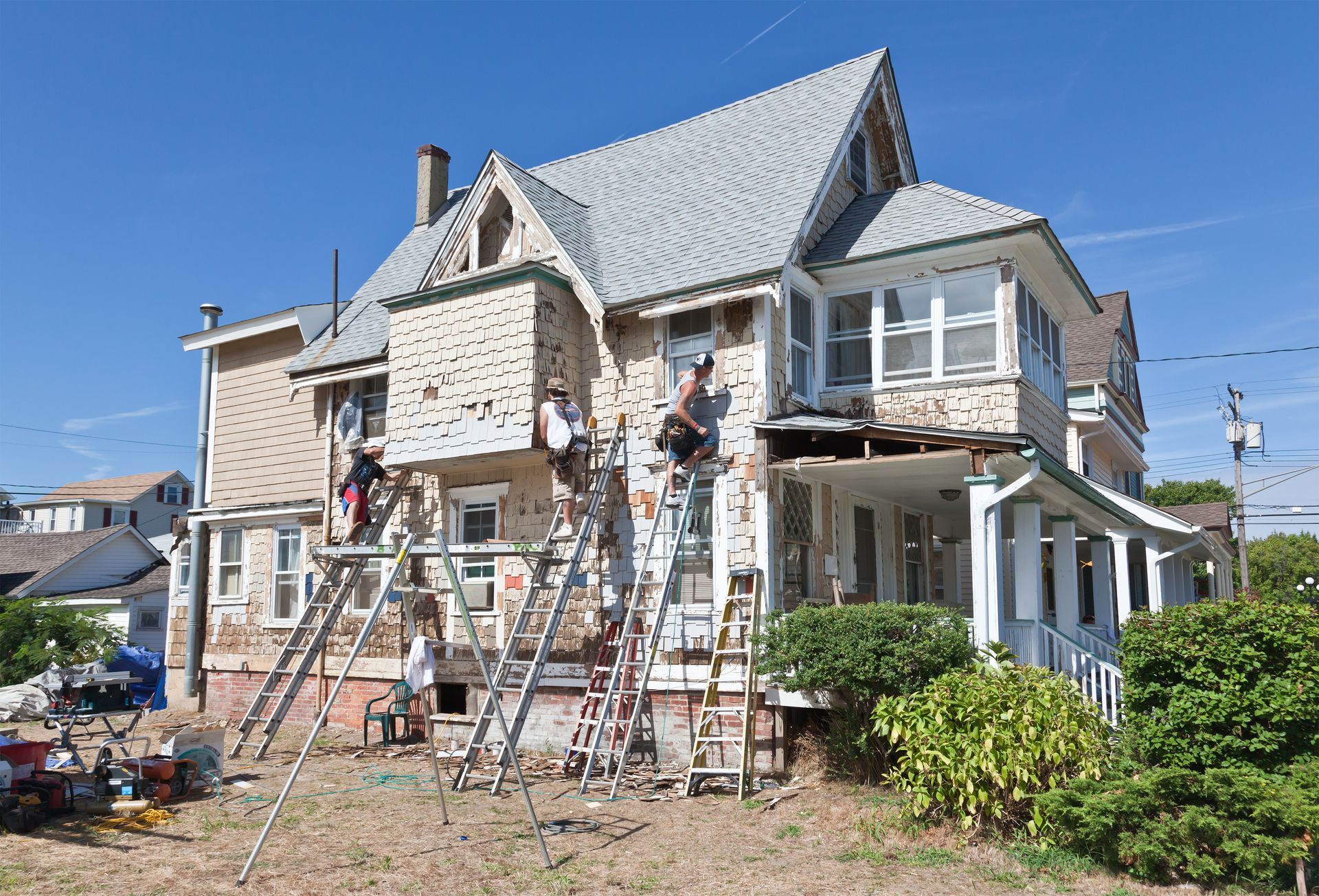 Workers on ladders repairing the exterior of a two-story house under a blue sky.