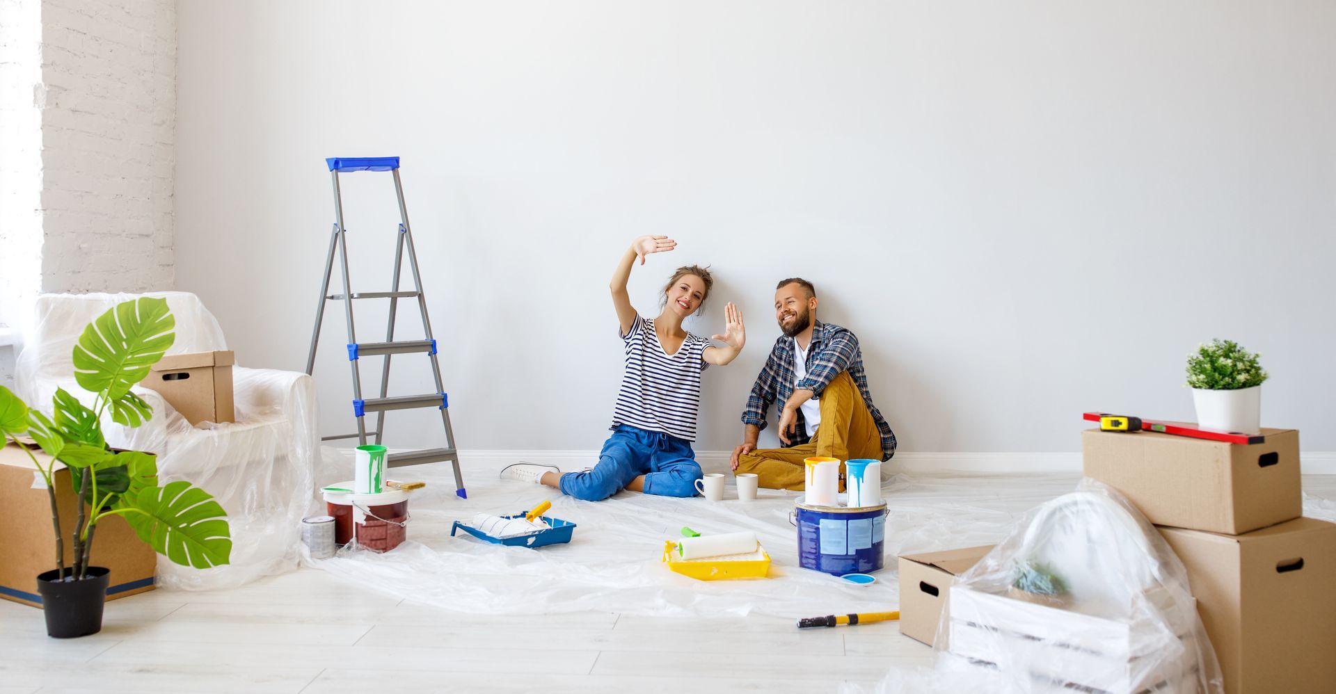 Two people painting a wall in a bright room during a home renovation, with ladder and supplies nearby