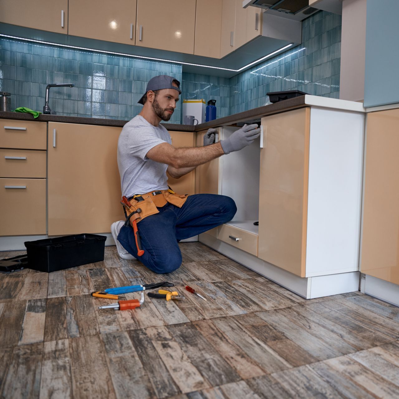a man is kneeling on the floor in a kitchen fixing a sink .