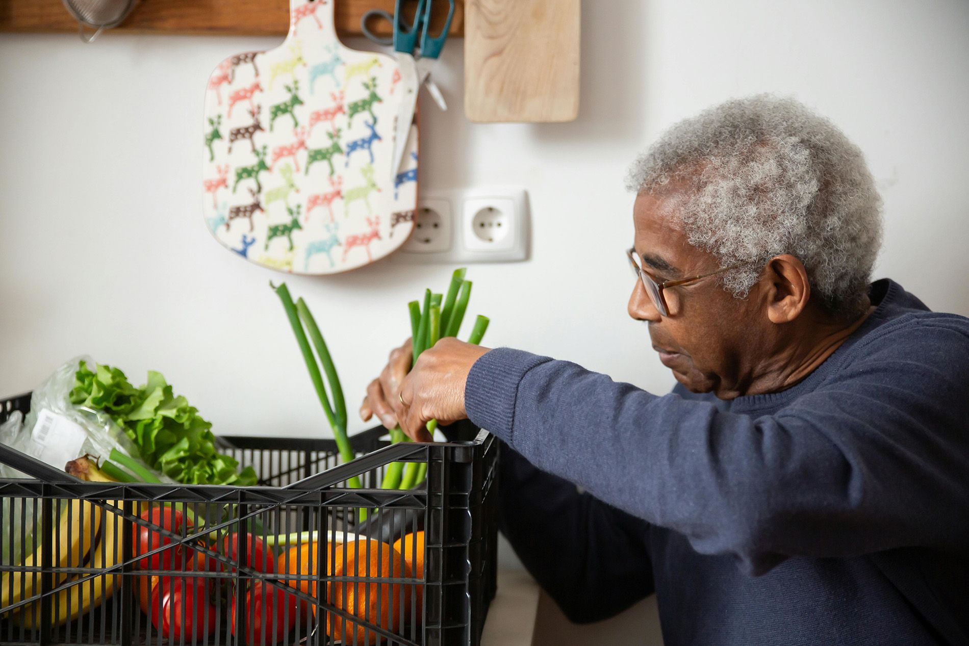 Older man in glasses sorting produce in a crate in a kitchen.