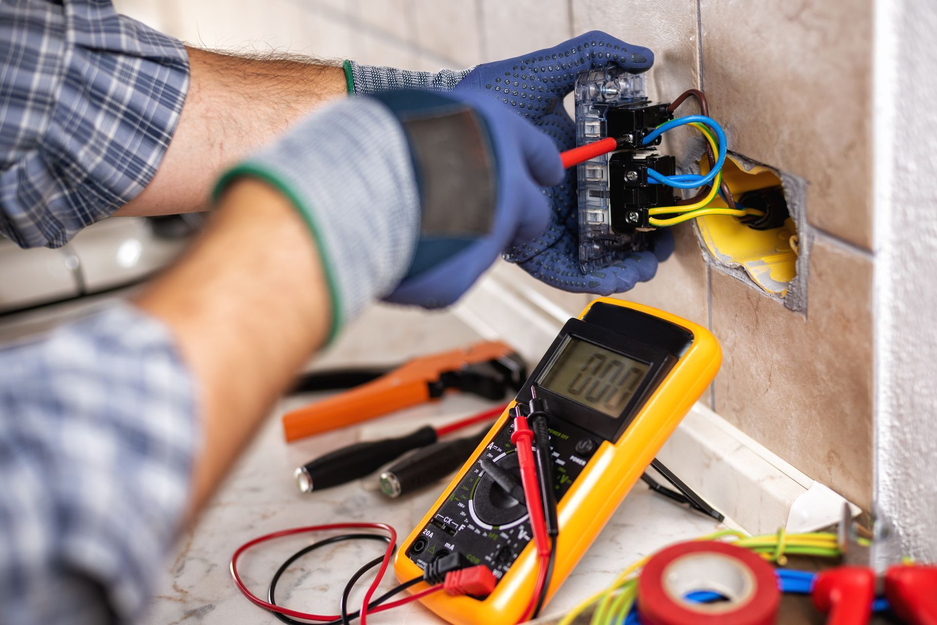 Electrician repairing wiring in a wall outlet using tools and a digital multimeter.
