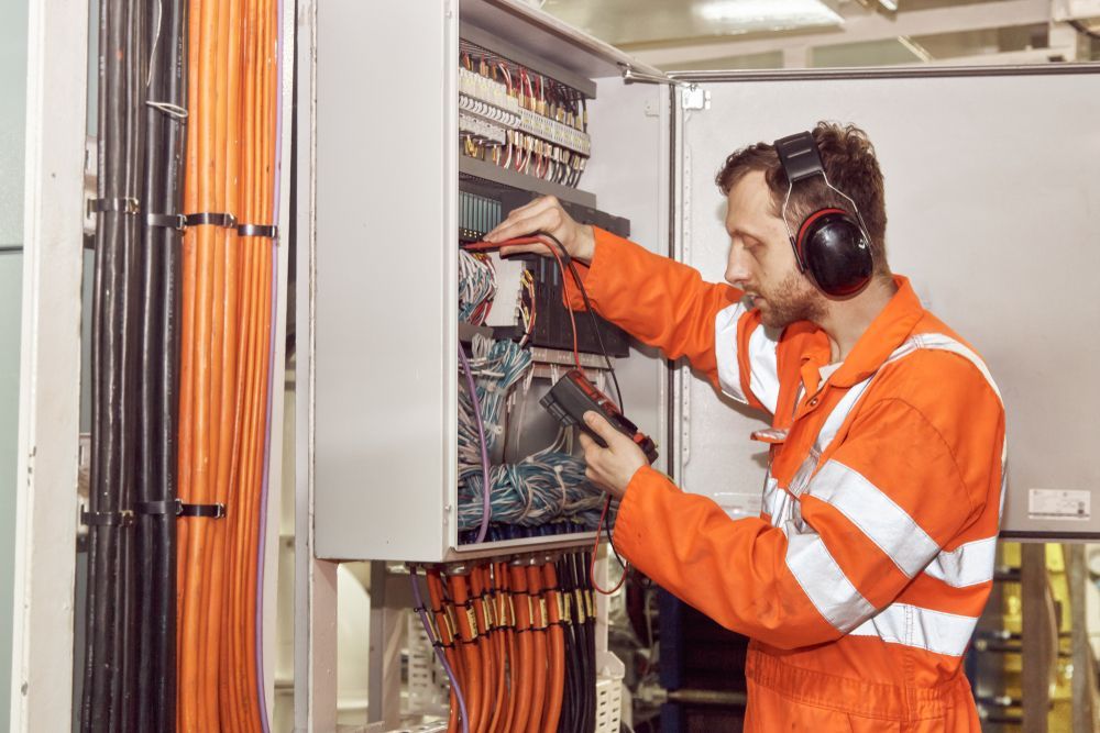 Electrician In Orange Overalls Tests Wires In An Electrical Panel — Quik Fire Electrical in Kelmscott, WA