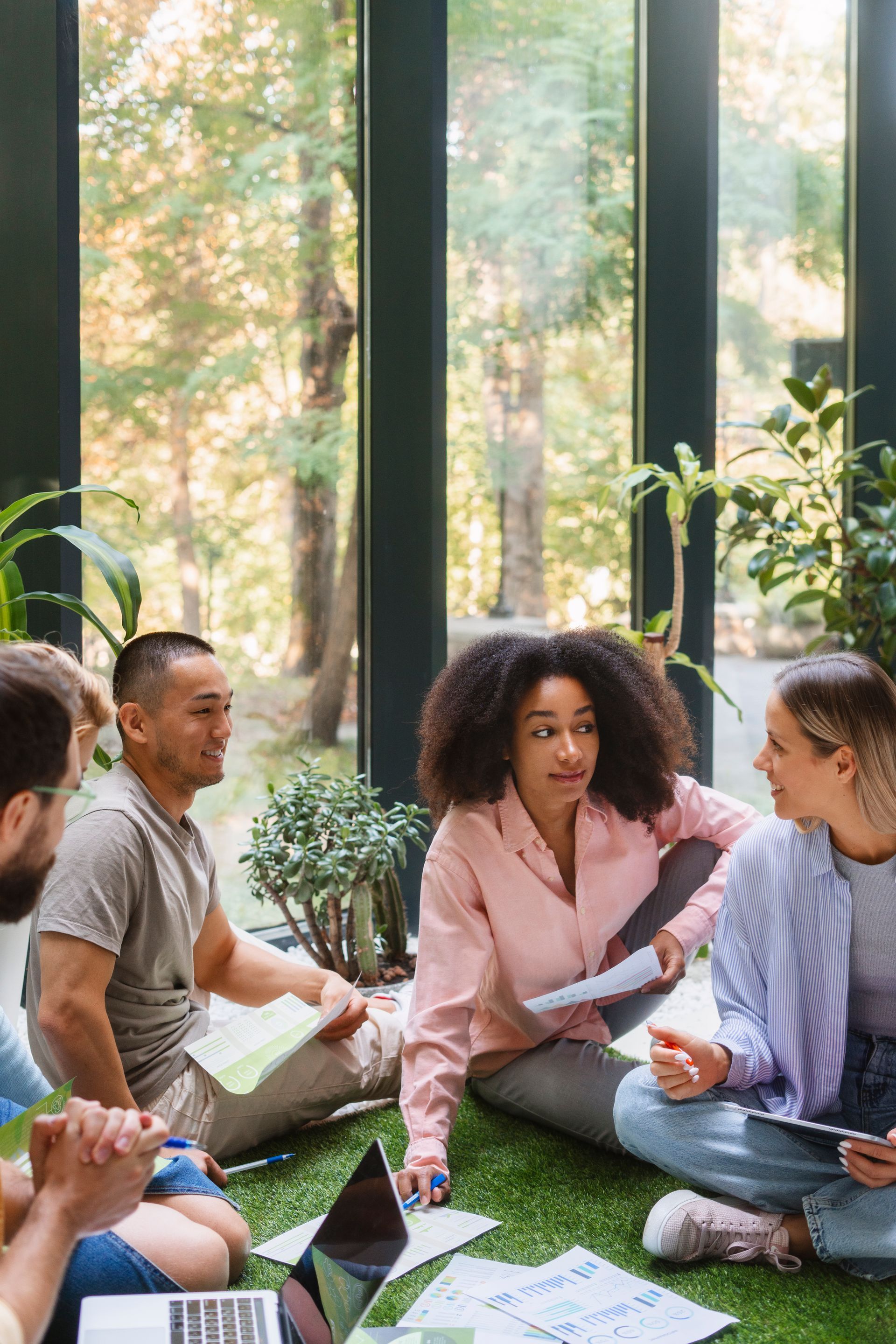 Groupe de personnes réunies sur une surface verte, discutant de documents. Bureau lumineux avec plantes et grandes fenêtres.