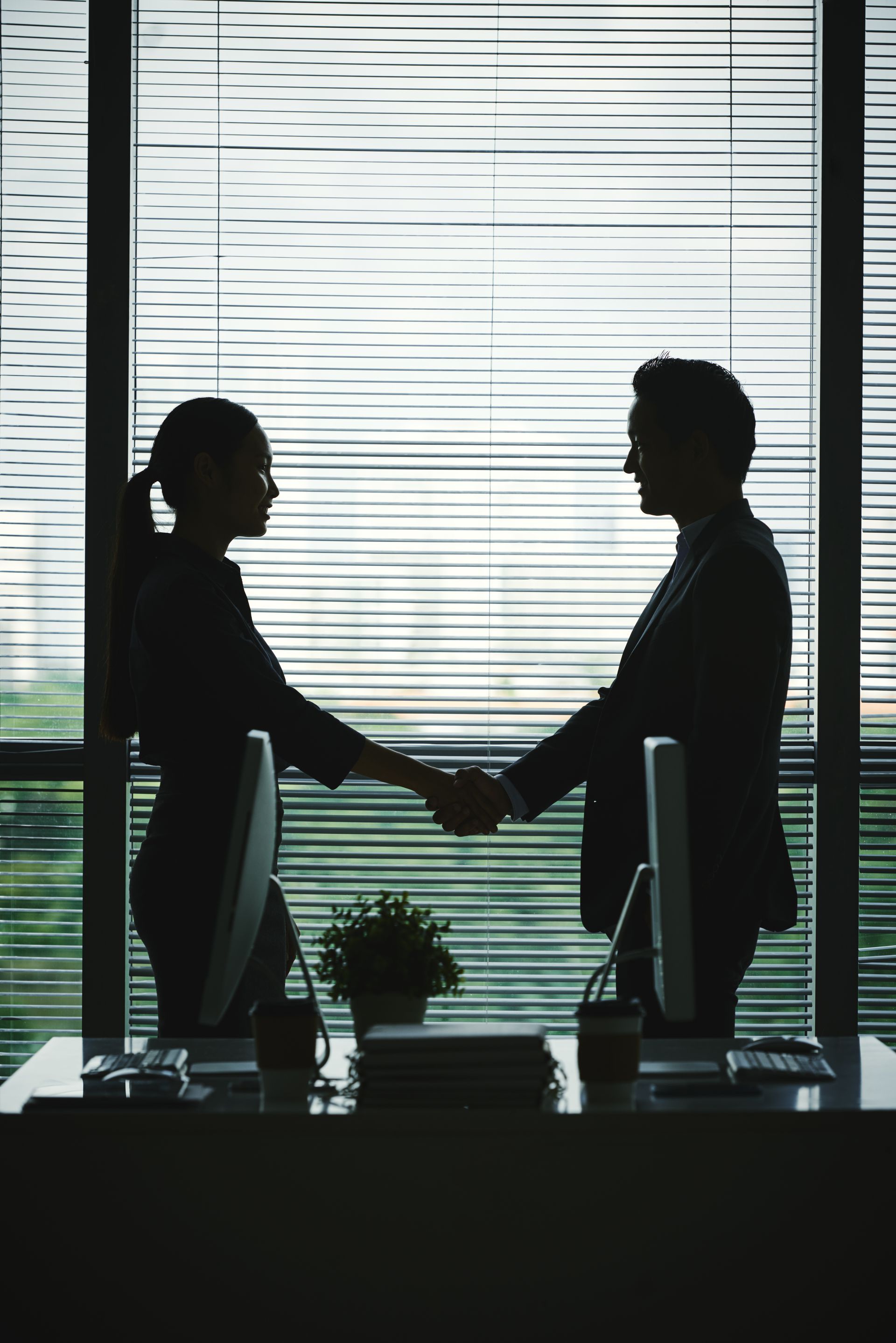 Deux personnes se serrant la main dans un bureau aux stores fermés, avec des ordinateurs de bureau.