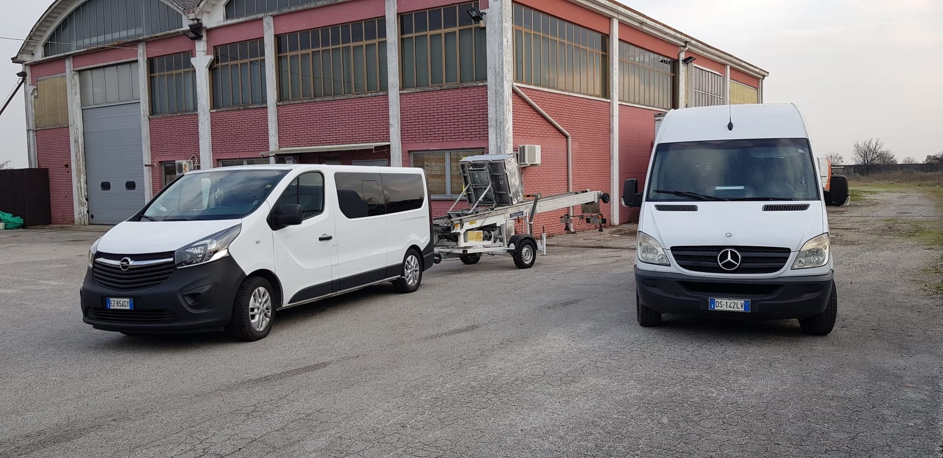 Tre furgoni bianchi parcheggiati sulla ghiaia vicino a un edificio rosso e grigio, con un piccolo carrello tra il furgone centrale e quello sinistro.