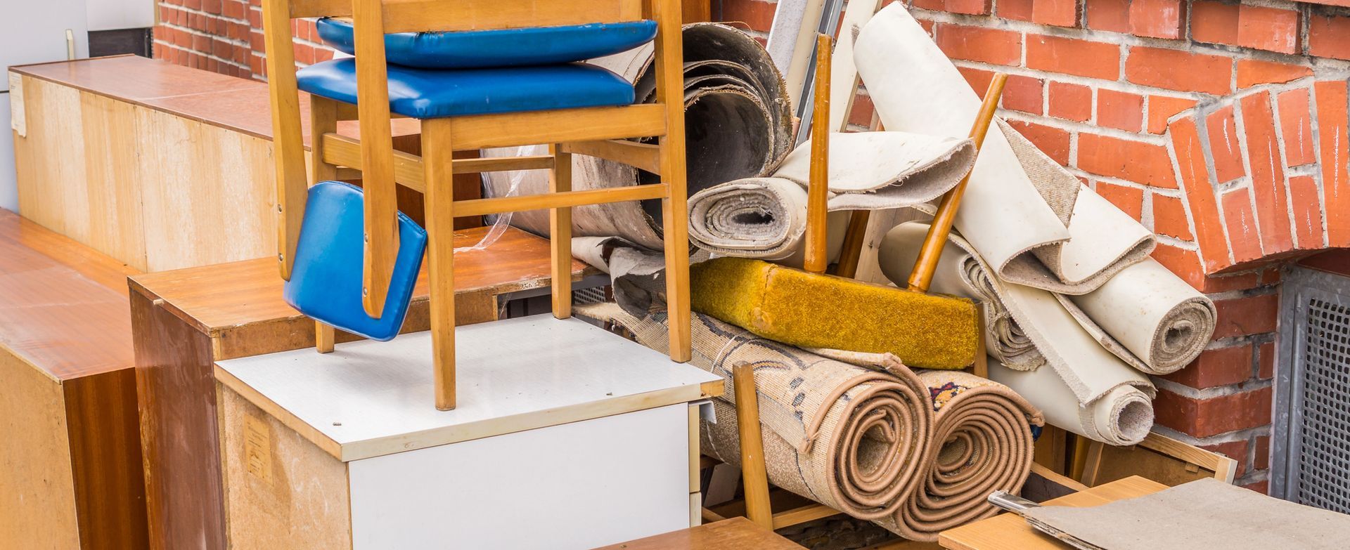 Oggetti ammucchiati, tra cui mobili, tappeti e una sedia, di fronte a un muro di mattoni.