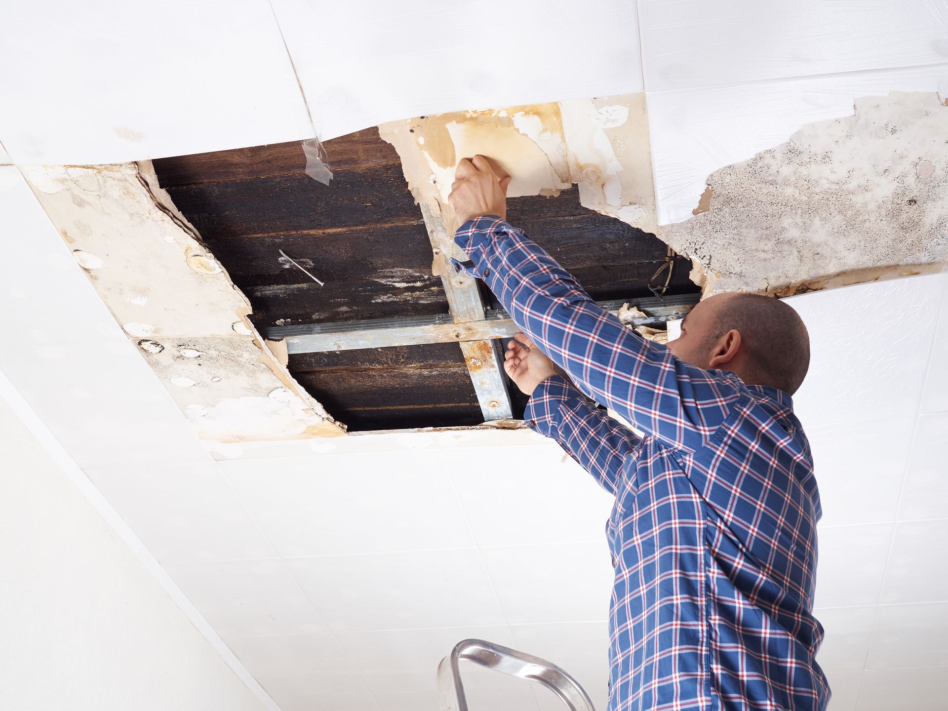 Man in a plaid shirt repairs a ceiling with a large hole. He is standing on a ladder.