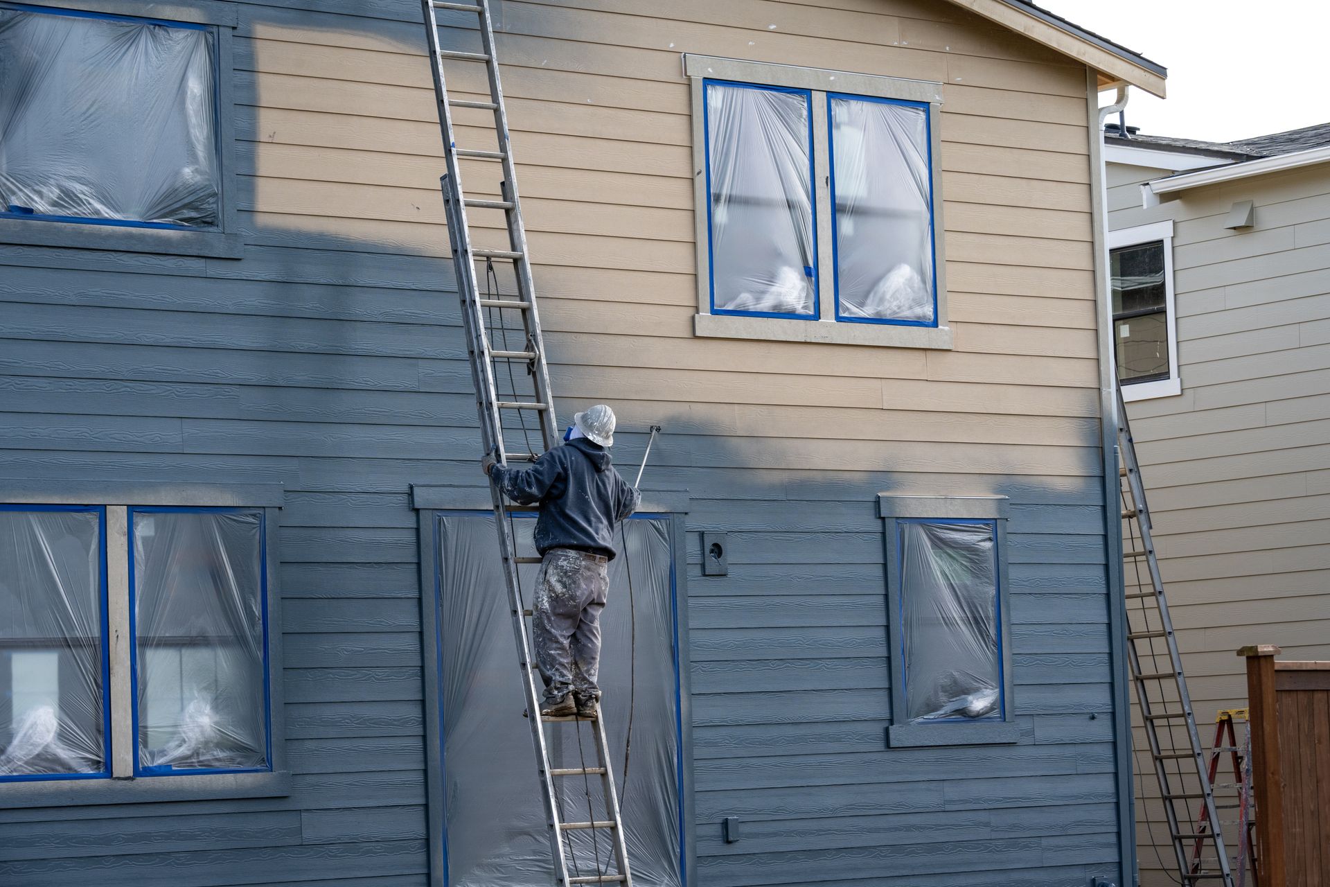 Person painting a house blue, standing on a ladder. Windows taped and covered with plastic.