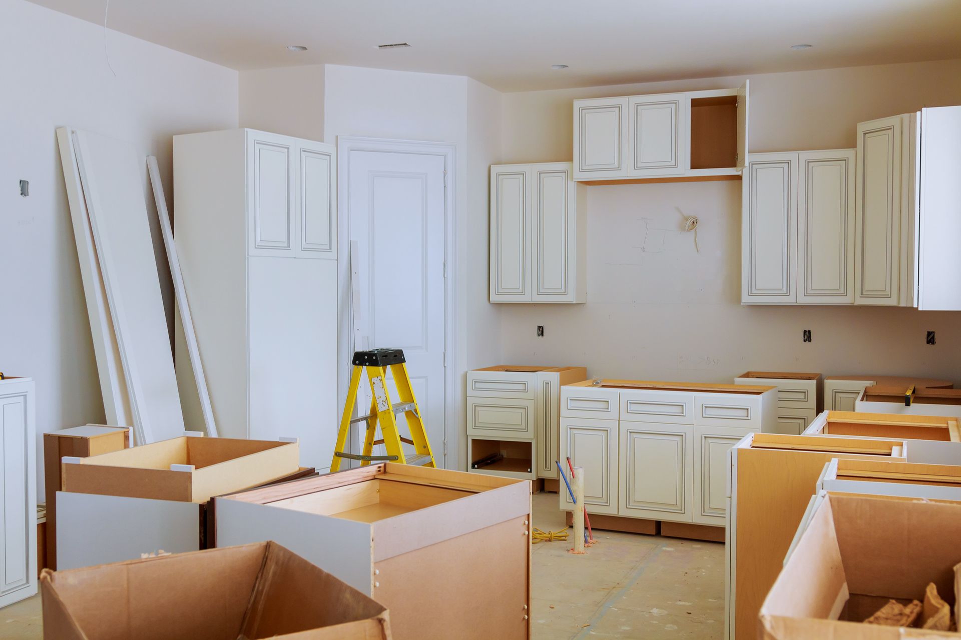 Kitchen under construction with white cabinets and cardboard boxes. Yellow ladder in the center.