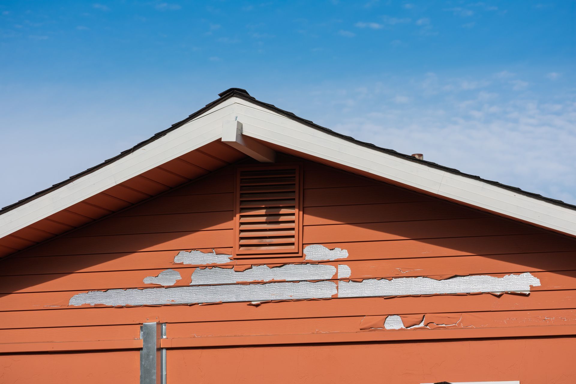 Orange house siding with peeling paint, vent, and white trim against a blue sky.