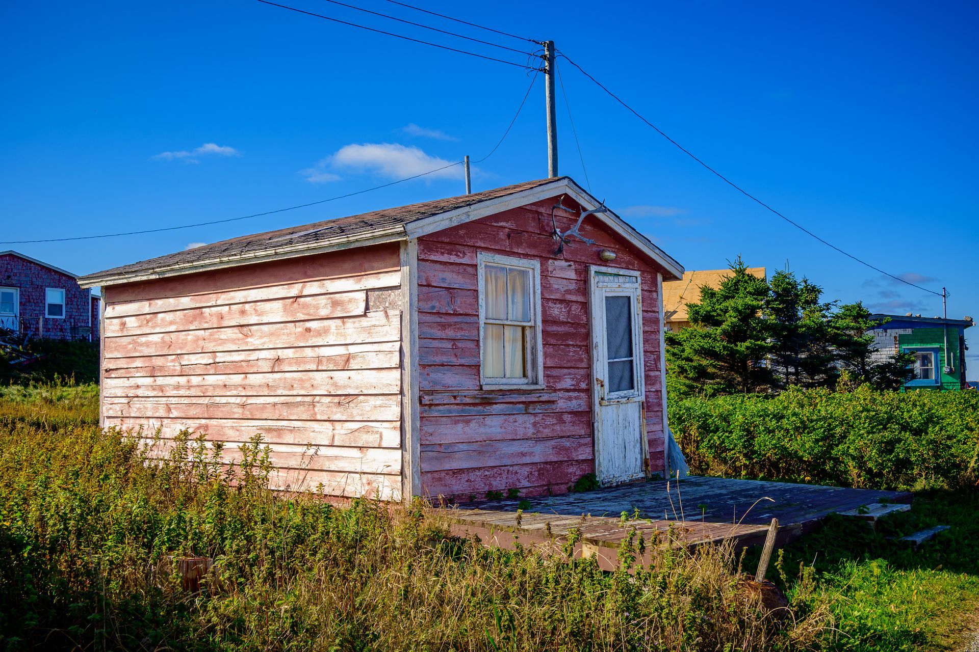 Red weathered shack with a white door and window sits on grassy land under a blue sky.