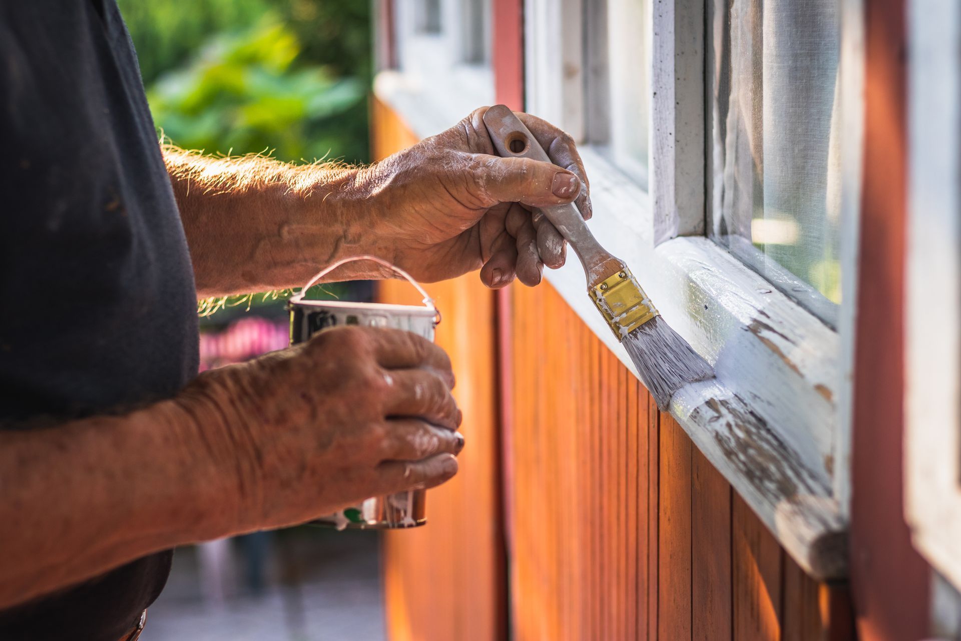 Person painting window trim with white paint, holding paint bucket. Red siding.
