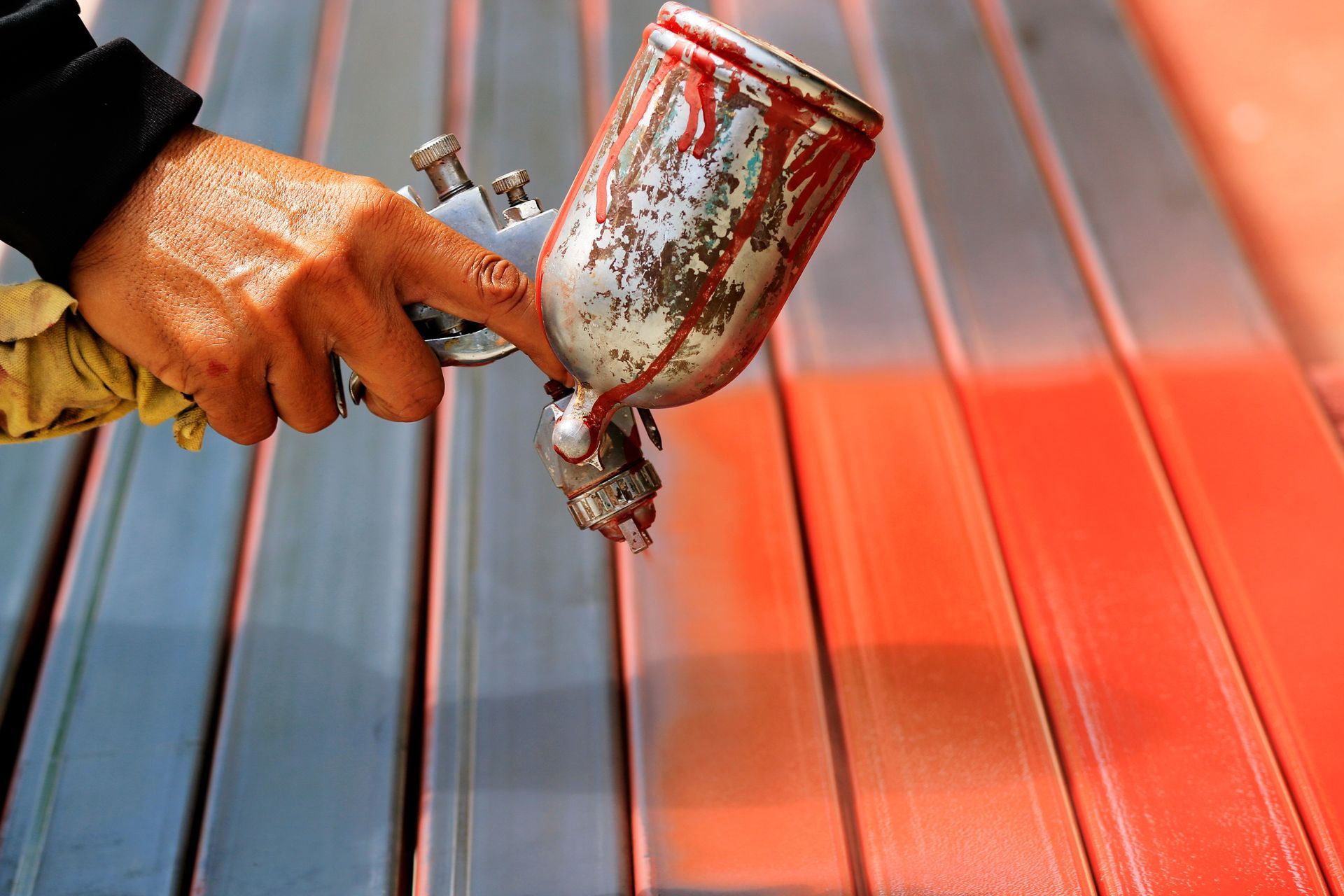 A hand sprays red paint on metal bars with a spray gun; half are painted red, the other half are gray.
