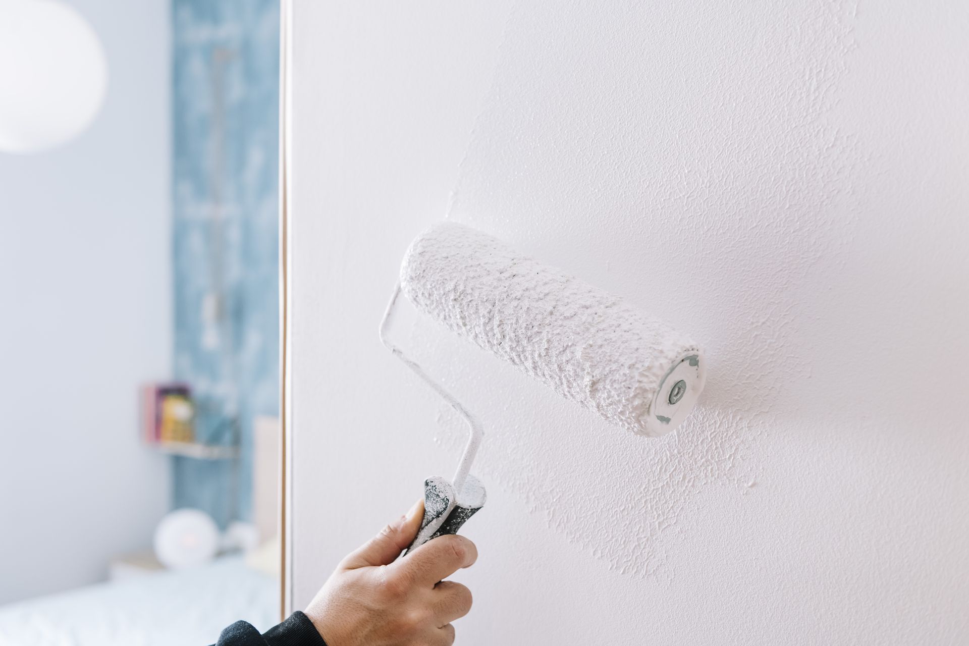 Person painting a white wall with a paint roller in a room.