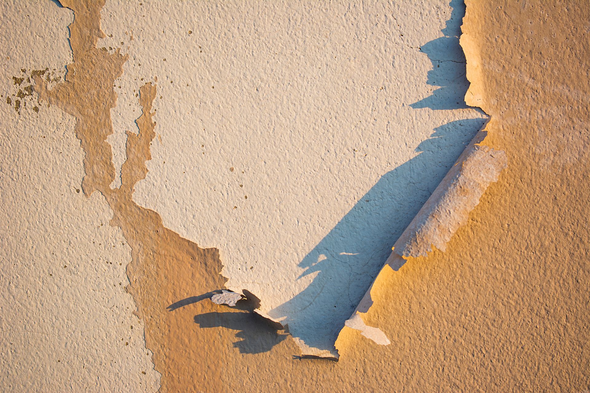 Peeling wall with beige and tan paint revealing a light blue layer. Shadow cast on the wall.