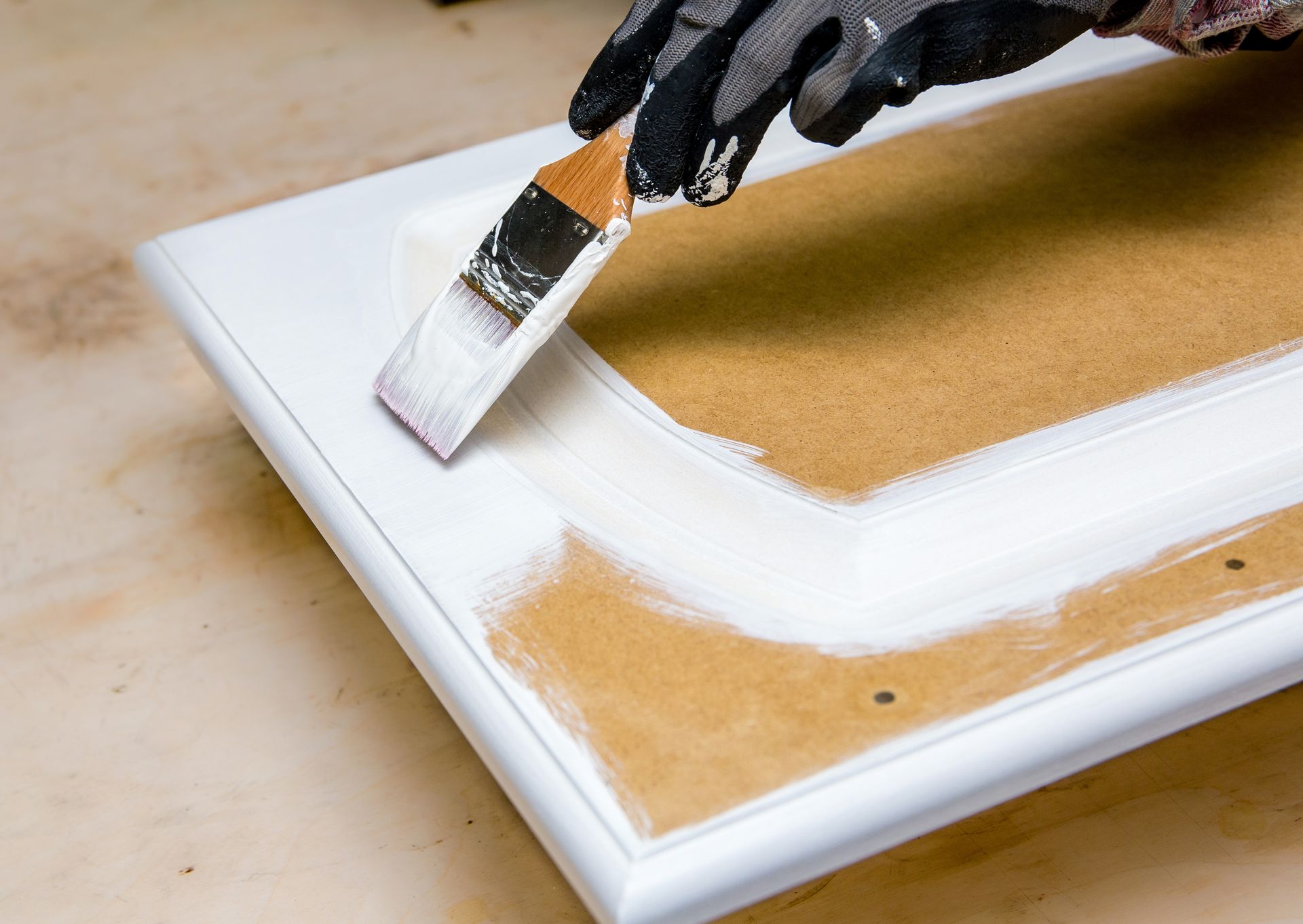 A person wearing a glove paints a white frame around a brown cabinet door with a brush.