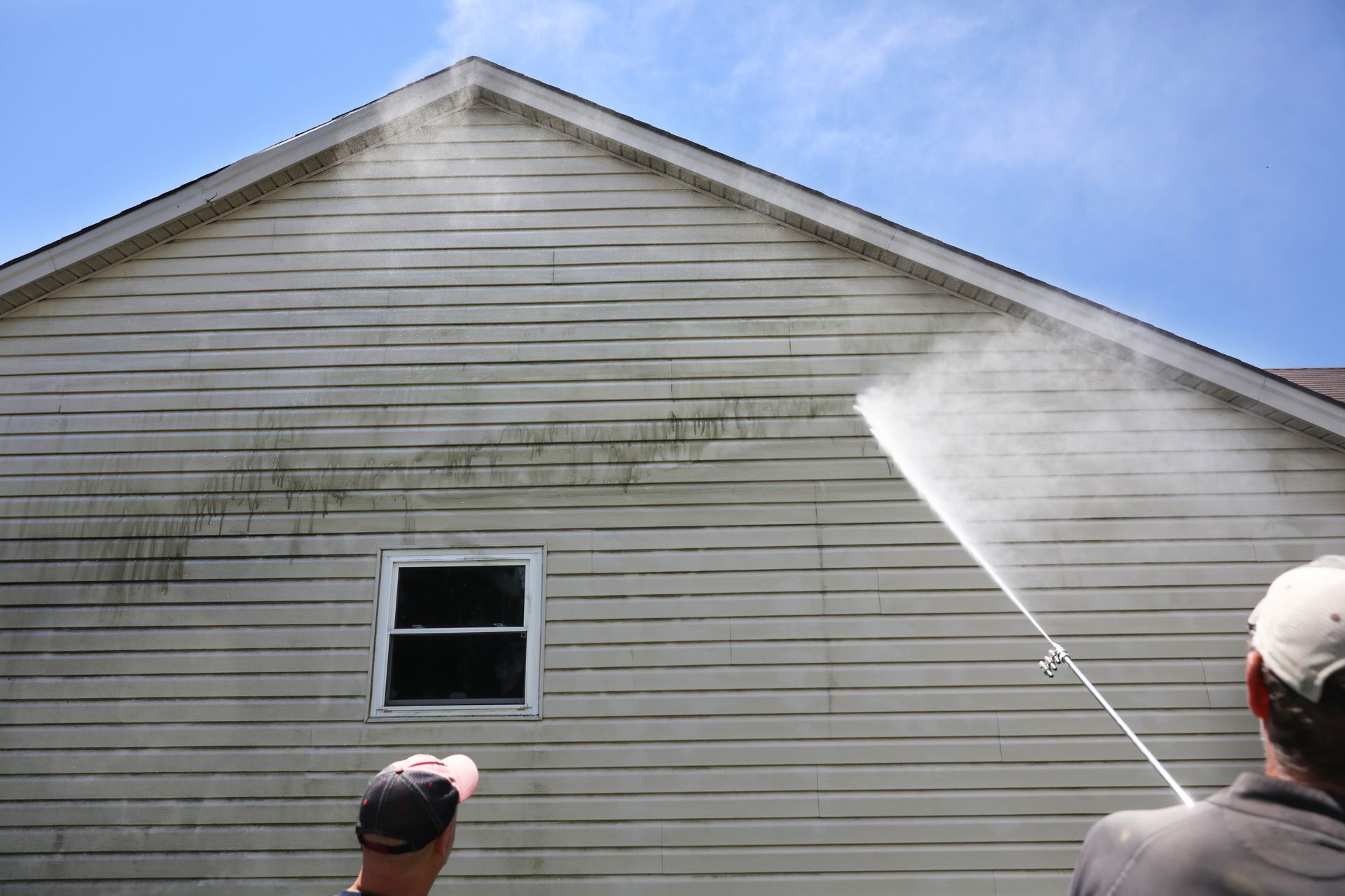 Man pressure washing the side of a house, removing grime and dirt, under a blue sky.