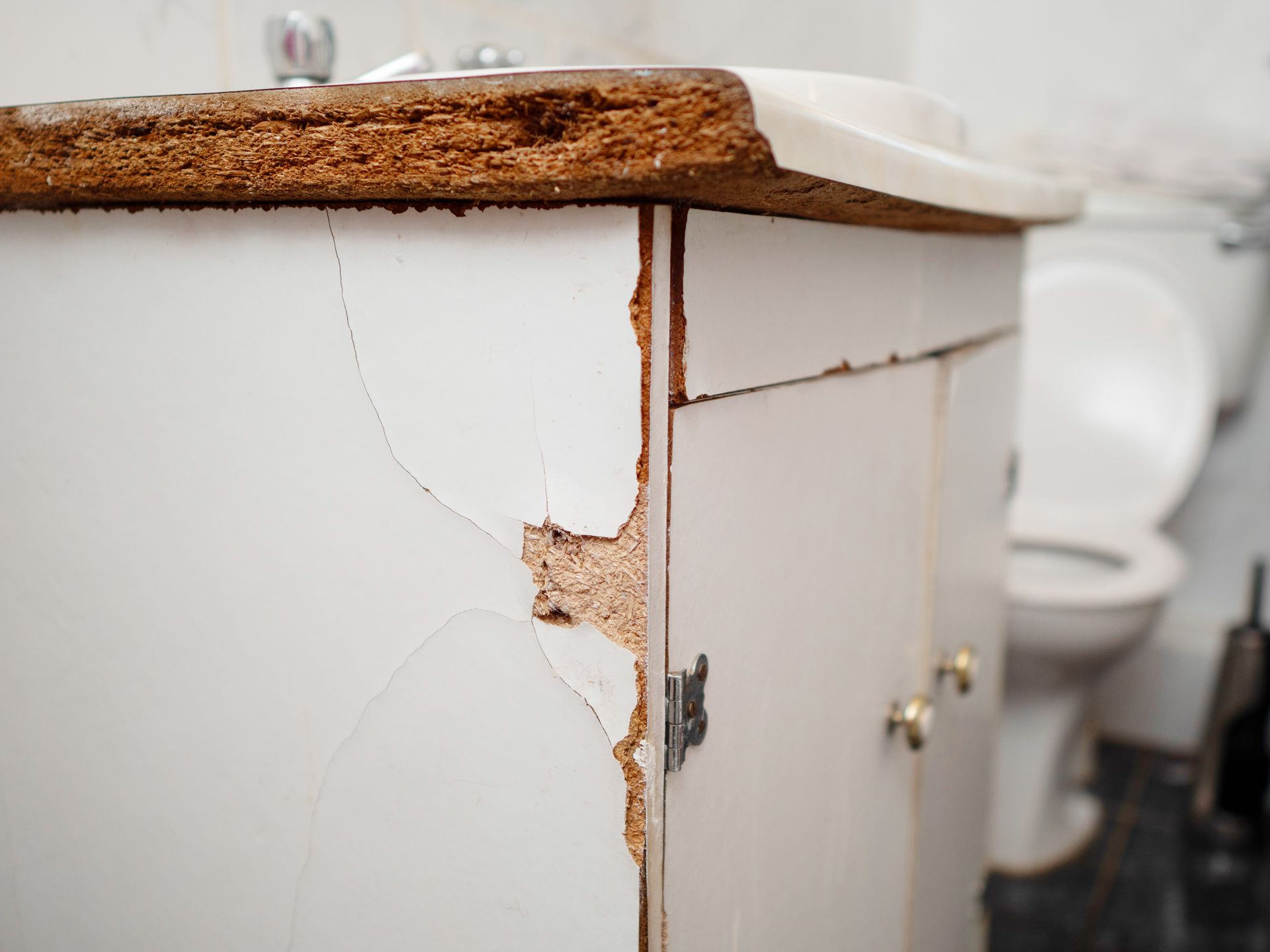 Damaged white bathroom vanity with cracked side, worn countertop, and toilet in the background.