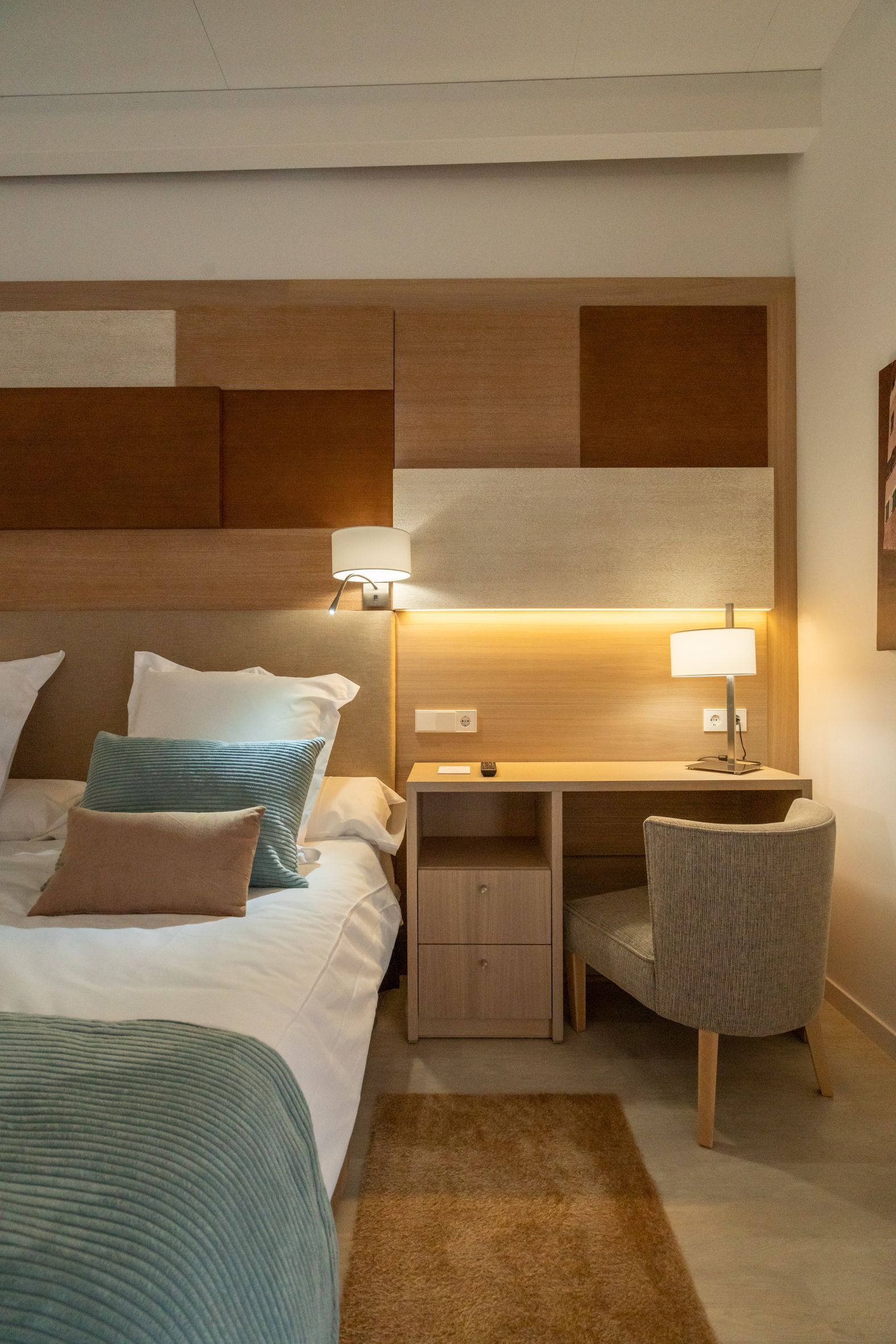 Bedroom with bed, desk, neutral tones, wood panel headboard, two lamps, textured chair, rug.