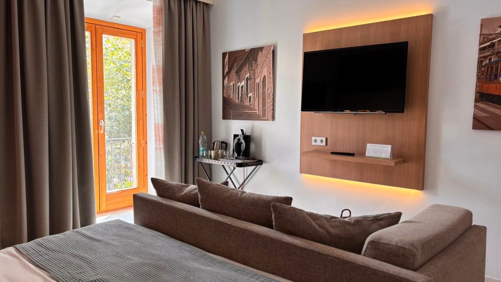 Cozy hotel room with a bed, sofa, and artwork. Warm lighting, neutral colors, and a window with curtains.