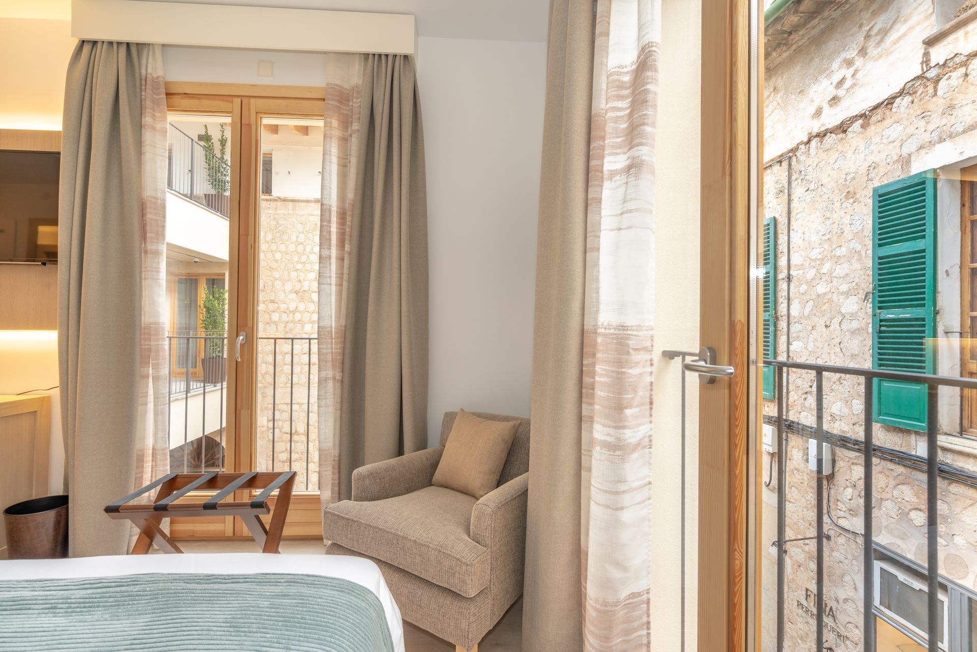 Room with balcony overlooking old building, tan curtains, armchair, luggage rack.