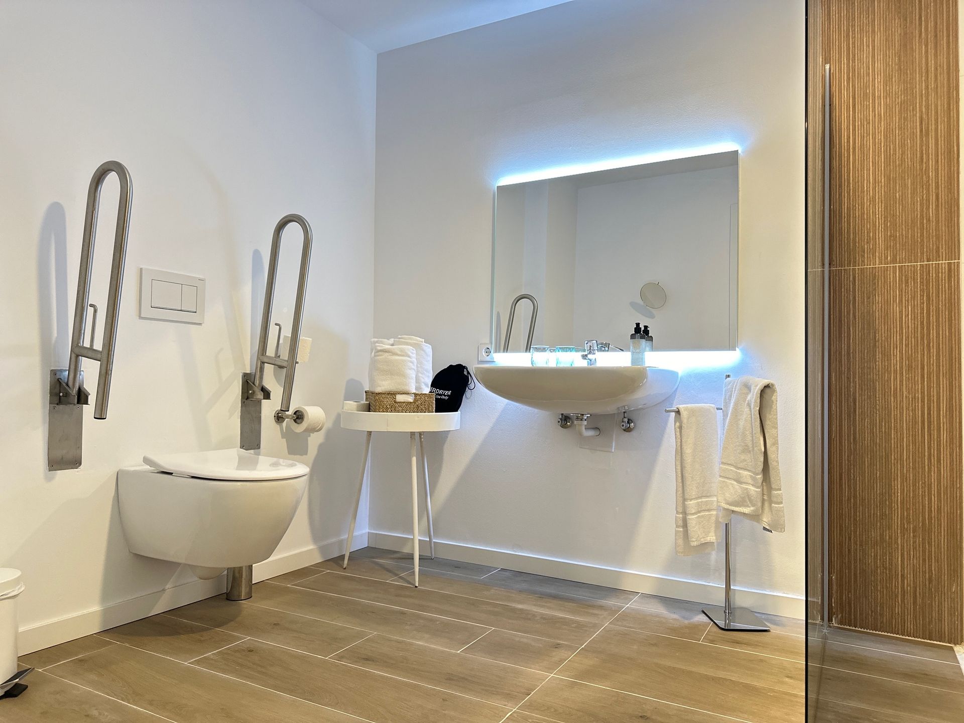 Barrierefreies Badezimmer mit schwebender Toilette, Waschbecken, Haltegriffen und beleuchtetem Spiegel.