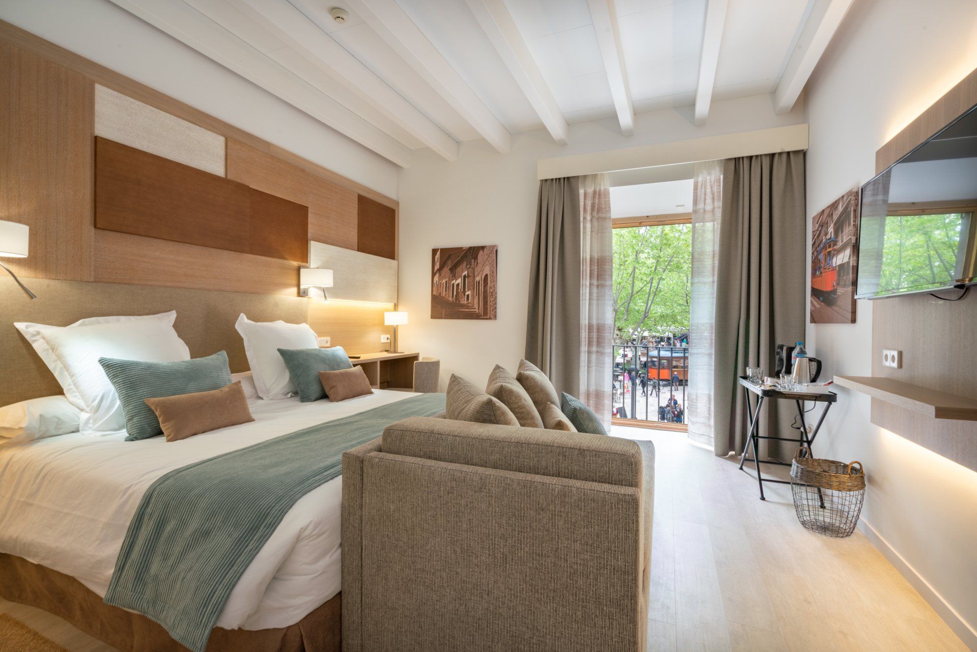 Chambre d'hôtel avec grand lit, canapé, balcon et télévision. Décoration beige et blanche avec touches de bois.