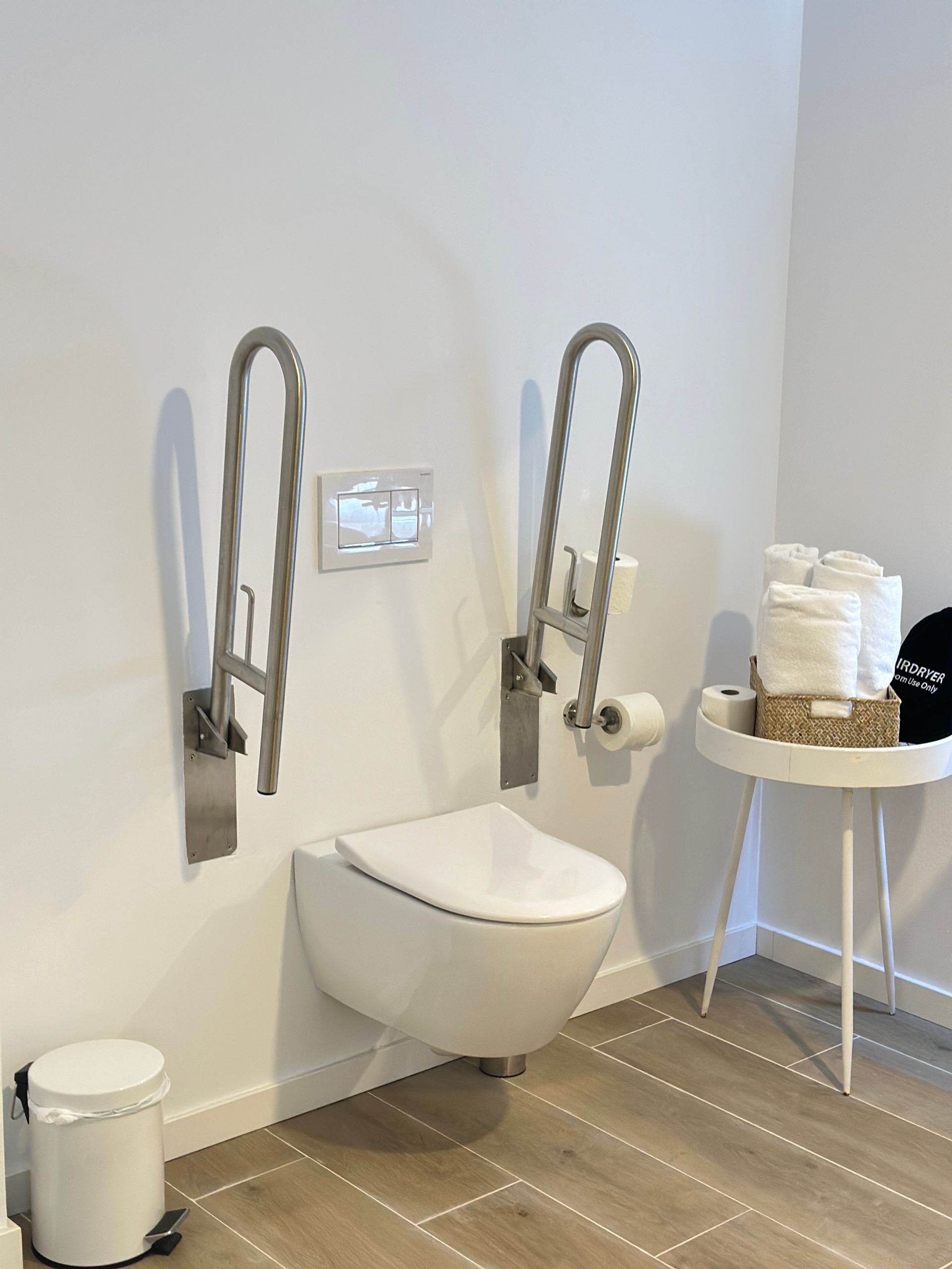 Baño accesible: inodoro con barras de apoyo, controles de descarga, papel, toallas en una mesa pequeña.