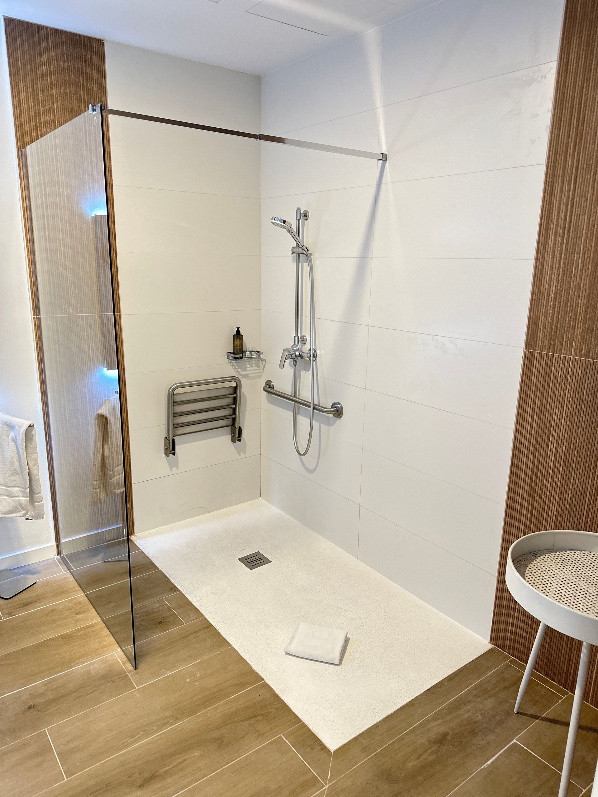 Douche accessible avec barres d'appui, siège intégré et cabine en verre, carrelage blanc et marron ; parquet en bois clair.