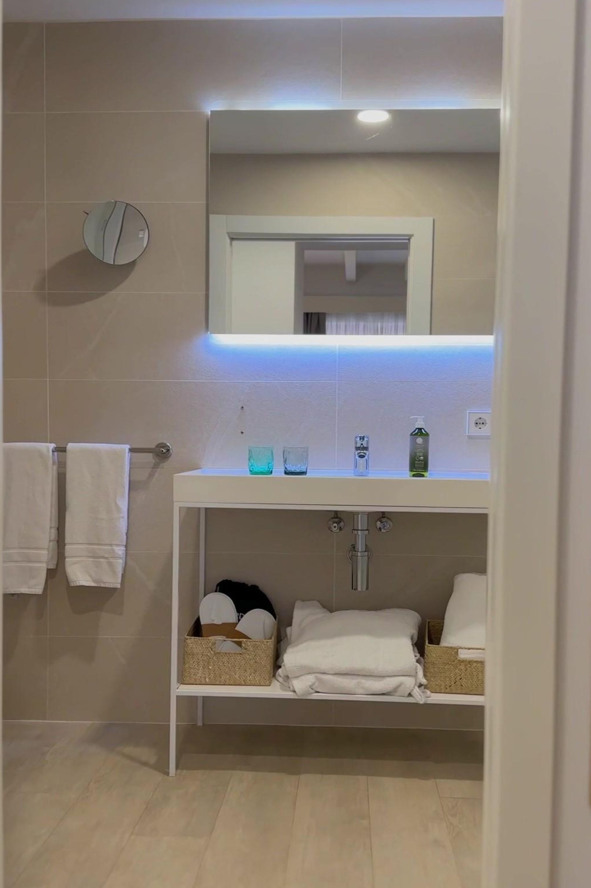 Bathroom with a white vanity, towels, mirror with blue backlighting, and beige walls and floor.