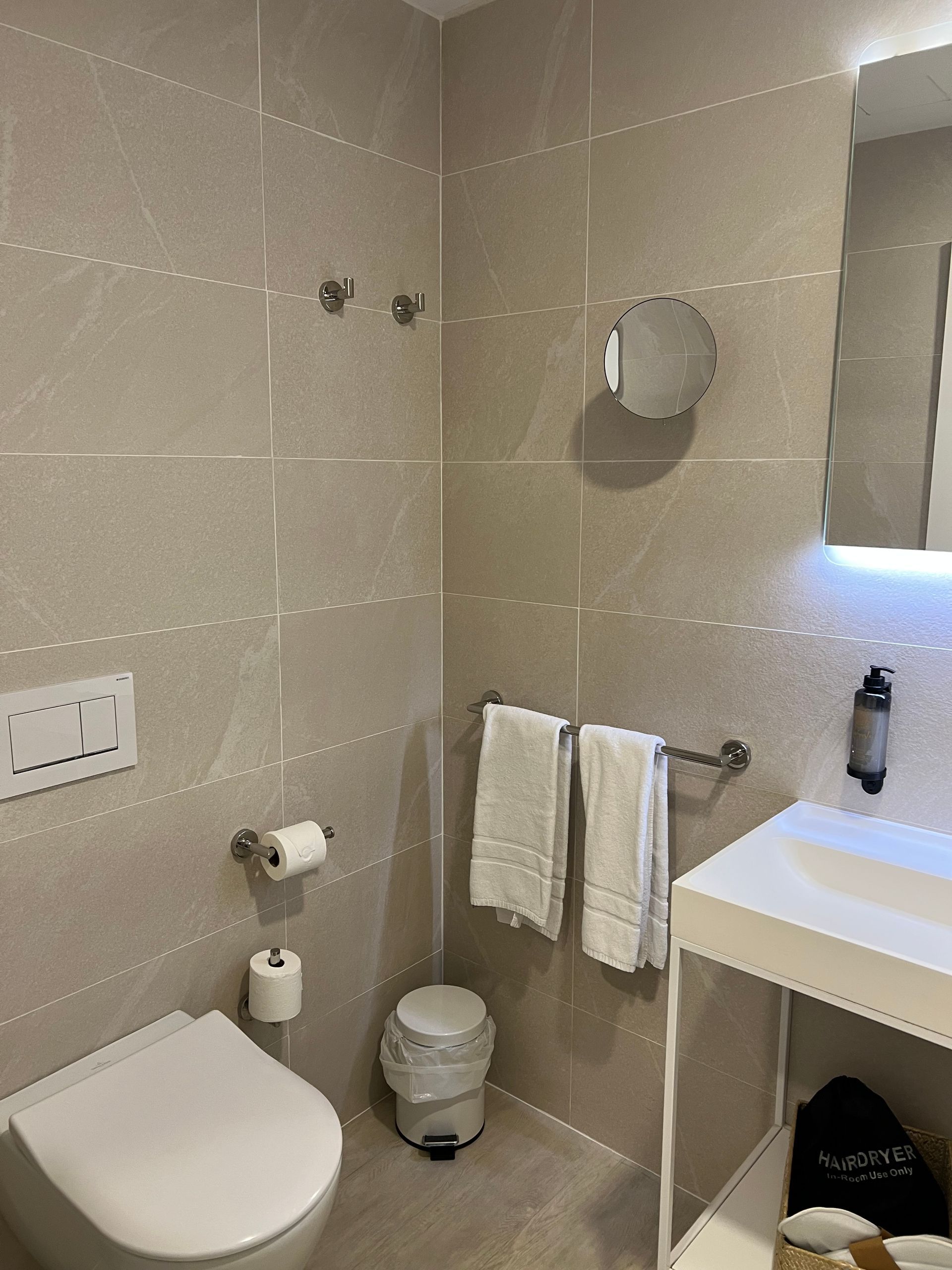 Baño con inodoro, lavabo, toallas y papelera. Paredes beige y grifería blanca.