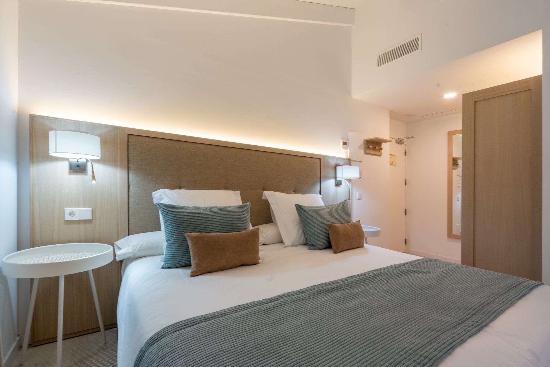 Bedroom with a bed, side table, and door. White bedding, blue and brown pillows, and light-colored walls.
