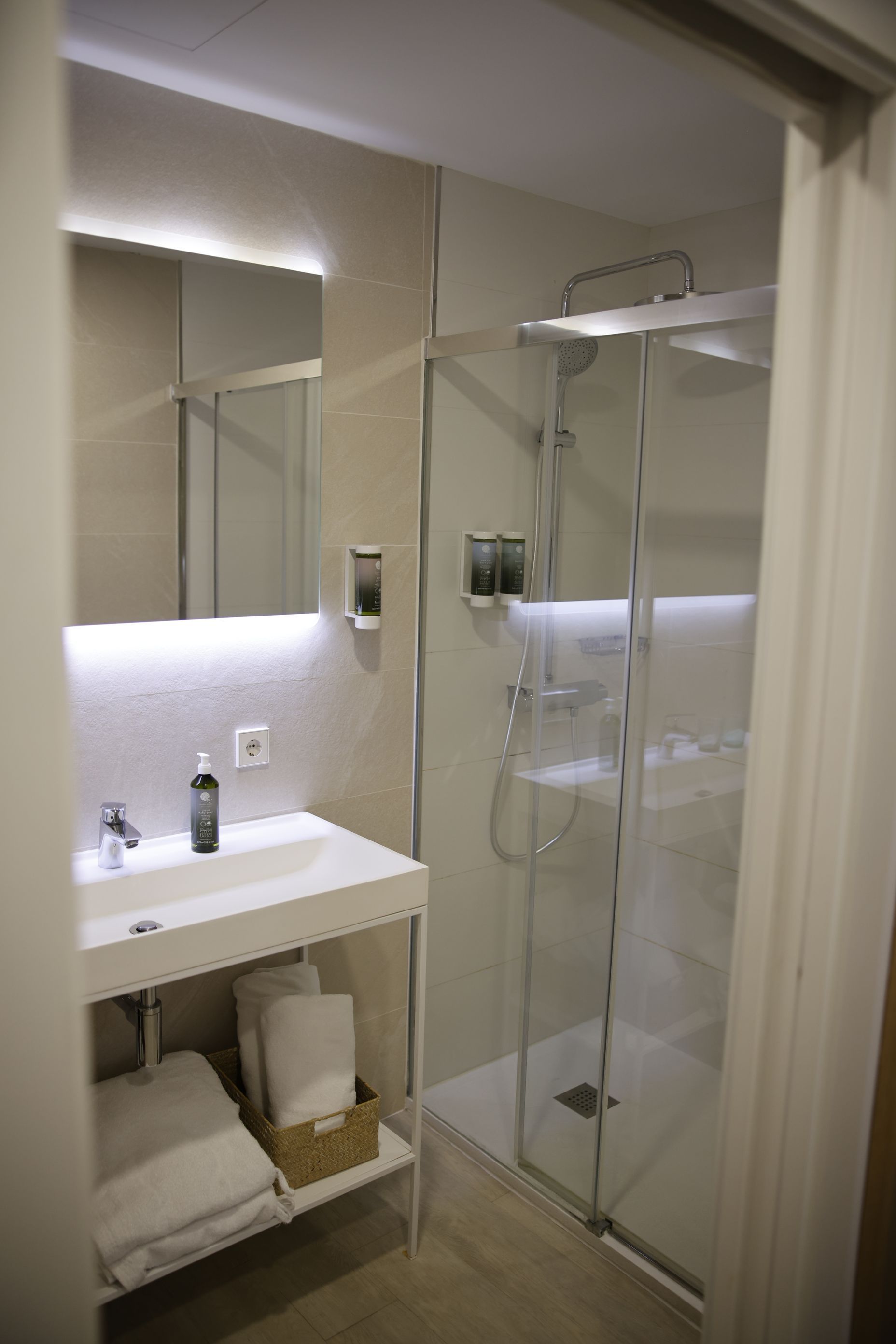 Baño moderno con ducha de cristal, lavabo, espejo con luces y toallas blancas.