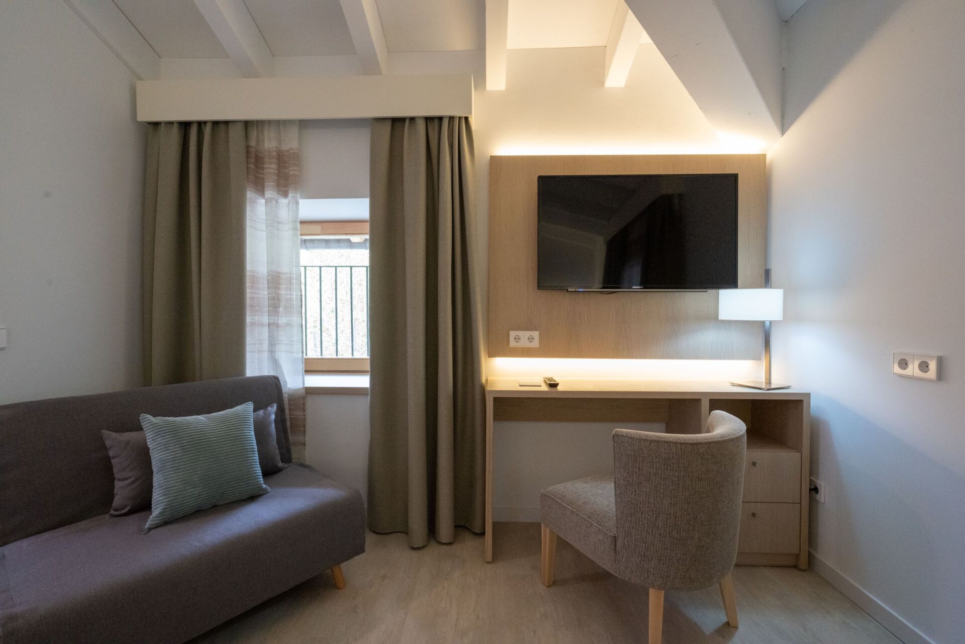 Ein Hotelzimmer mit Sofa, Schreibtisch, Fernseher und einem mit Vorhängen bedeckten Fenster.