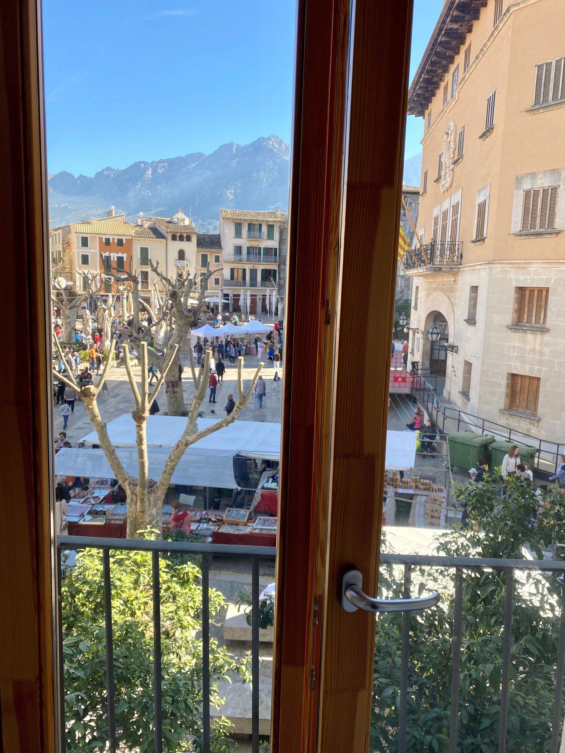 View from a window: a plaza with market stalls, buildings, and distant mountains under a blue sky.