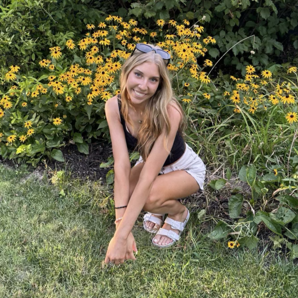Young caucasian woman bent down next to yellow flowers in white shorts, black tank top, and white sandals. Blonde long hair. 
